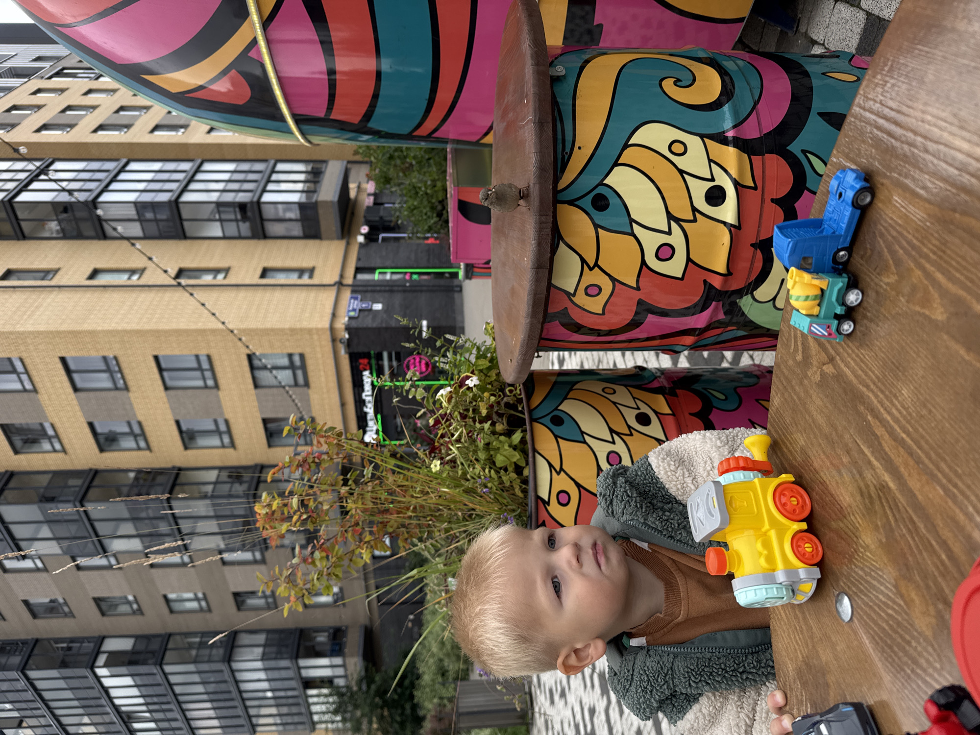 In Novoselye, Russia, a blonde-haired toddler with a serious expression sits at a wooden table. He wears a green fuzzy jacket over a brown shirt. On the table are several toy vehicles: a yellow train with red wheels and orange accents, a blue and green cement mixer truck, and parts of other toy cars. A small sparrow stands on the table near the toddler, facing away from the camera, its beak slightly open.
The background features colorful, cylindrical art installations and modern apartment buildings. One installation, directly behind the toddler, is decorated with a yellow, orange, and red floral pattern on a teal background. Another, to the right, displays a vibrant, abstract design with pink, orange, yellow, and teal elements. The buildings in the background are a mix of beige brick and darker-toned facades with numerous windows. A street lamp string with visible bulbs is strung between the buildings. The ground is paved with a light-colored patterned surface.