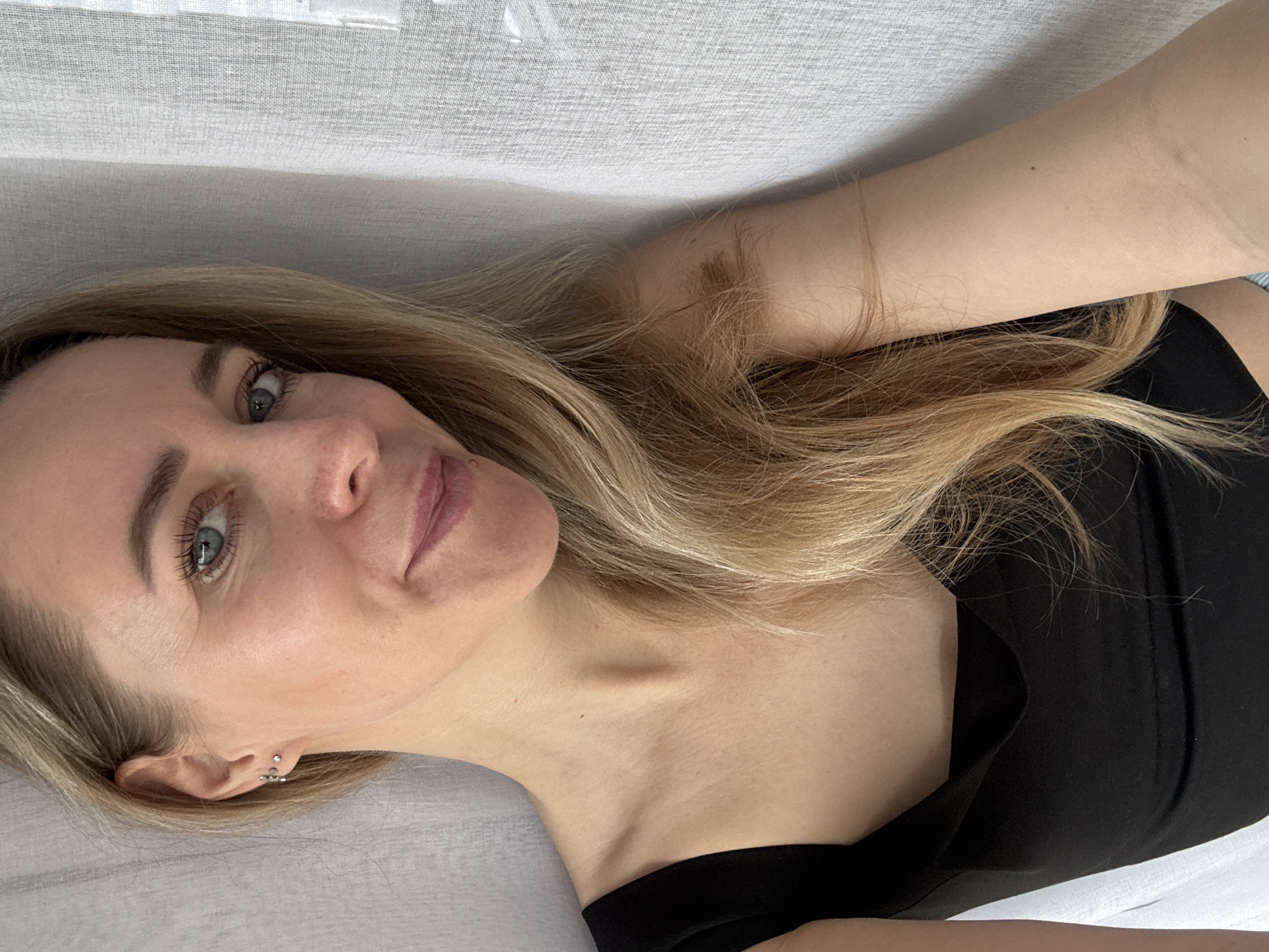 A woman with light blonde hair and blue eyes is lying down on a white surface. She is wearing a black top. She has multiple piercings in her left ear. The background appears to be a white curtain or draped fabric. The location is not identifiable as Chaikovsky, Russia, from the visual information provided.