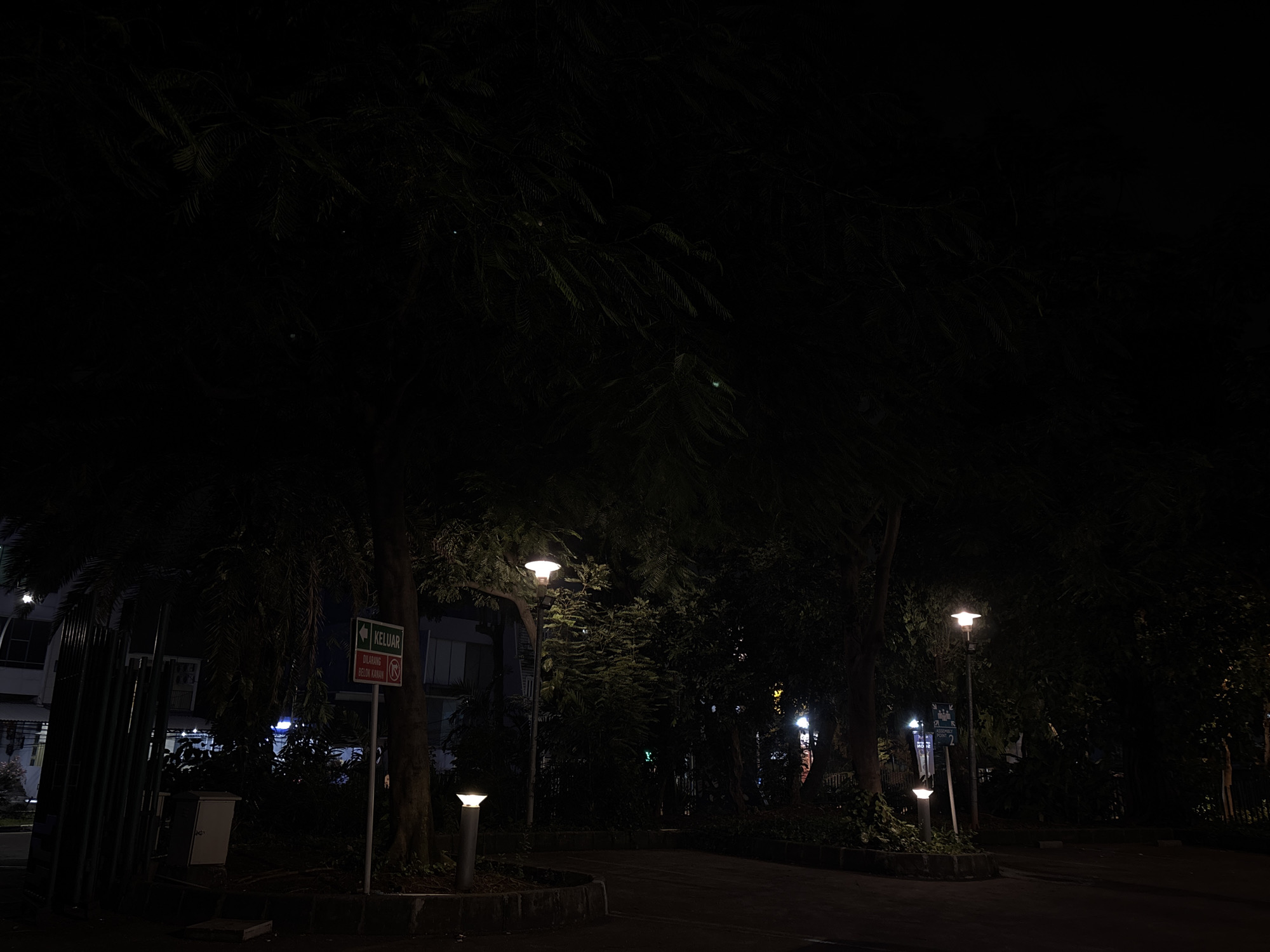 The image captures a night scene in North Jakarta, Indonesia. The foreground consists of a paved area with street lamps providing illumination. Several trees with dense foliage dominate the view, casting shadows and obscuring much of the background. On the left, a sign with the Indonesian word "KELUAR" (Exit) and another sign prohibiting right turns is visible near a building. The surrounding environment appears to be a public park or a residential area. There are no people visible in the image.