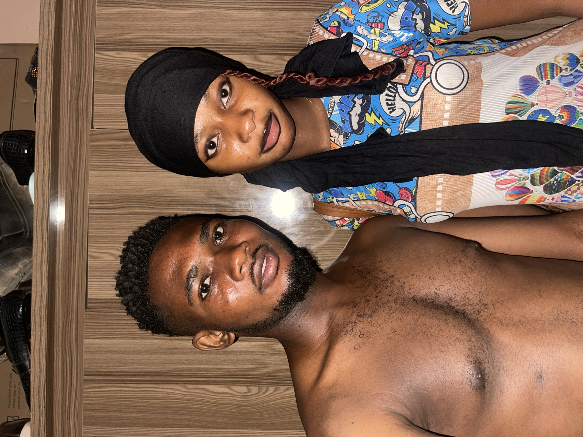 The image shows two dark-skinned individuals on a wooden surface, possibly in Muchiya, Nigeria. The individual on the left is a man with short, dark hair and a beard, shirtless, gazing towards the camera. To the upper right is a woman wearing a black head covering and a colorful patterned shirt with cartoonish images, also looking toward the camera. Part of a mirror and some objects are barely visible on the left side. The wooden surface appears to be a floor or a wall.