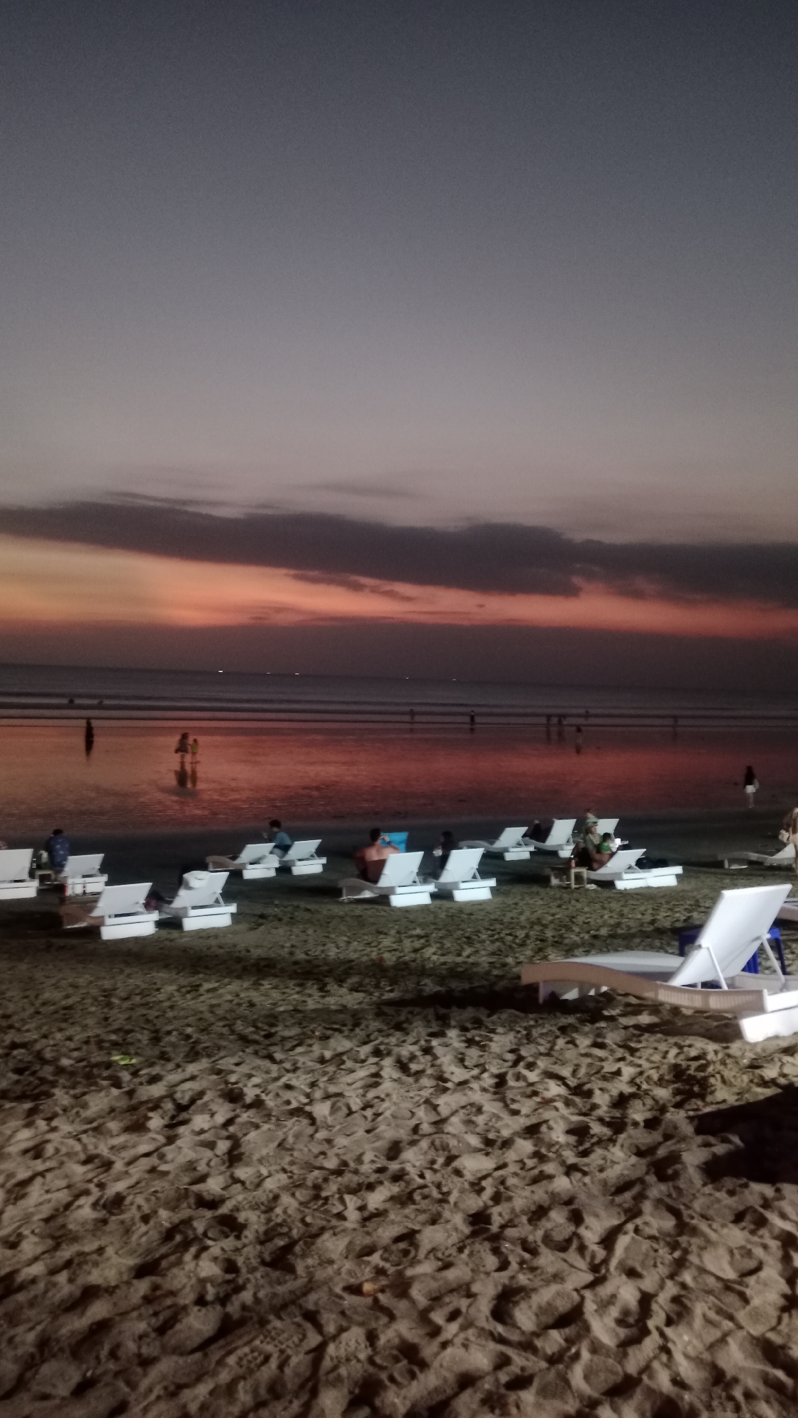 This is a wide shot of a beach in Kuta, Indonesia, at dusk. The sky is a gradient of dark grey at the top to a soft pink and orange near the horizon. The water is calm and reflects the colors of the sky. Several people are visible in the shallow water near the shore.
In the foreground, the sandy beach is covered with several white lounge chairs, most of them empty. A few people are seated in some of the chairs. The sand is textured with footprints and small indentations. The lighting suggests it is twilight, with some artificial lights from the beach area likely contributing to the illumination. In the background, the ocean stretches out to the horizon, where a few small lights are visible. The overall atmosphere is serene and relaxing, indicative of a beach resort in Kuta.