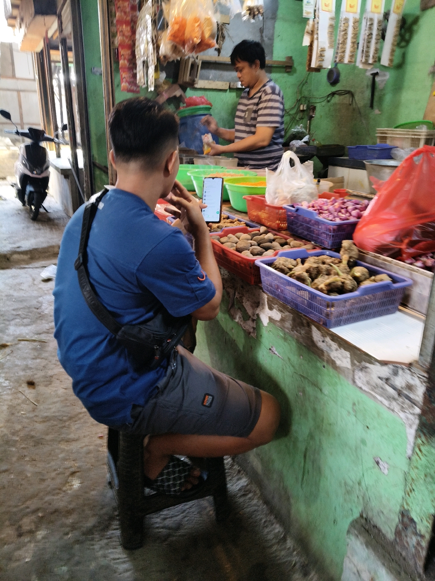 The image depicts a market stall in Setiamekar, Indonesia. A young man, wearing a blue t-shirt and grey shorts, sits on a dark stool in the foreground, his back to the camera. He has a black sling bag across his chest and is looking at a smartphone in his hand. Behind him, a vendor in a striped t-shirt stands at the counter, preparing goods. The stall is laden with various items, including baskets of shallots and root vegetables. A red plastic bag is visible on the right side of the counter. In the background, a scooter is parked near the entrance of the stall. The stall's walls are painted green and appear weathered.