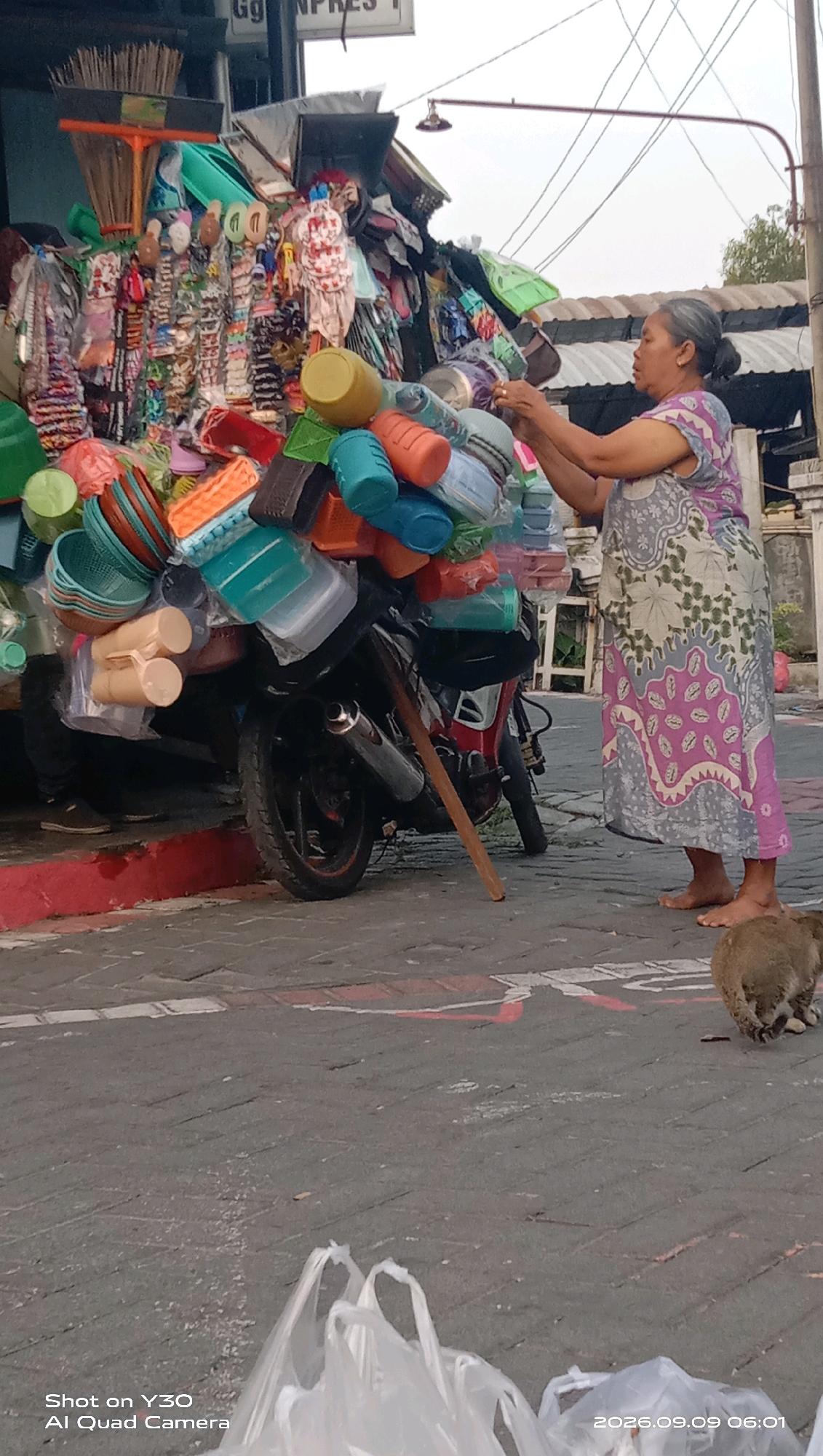 A woman in a patterned dress is standing next to a motorcycle loaded with various plastic containers and household items. The motorcycle is parked on a paved street in Klangonan, Indonesia. The woman is holding onto the stacked items, appearing to adjust or organize them. A small animal, possibly a cat or a dog, is walking away from the camera in the foreground. In the background, there are market stalls and buildings, indicating a commercial area. The sky is overcast.