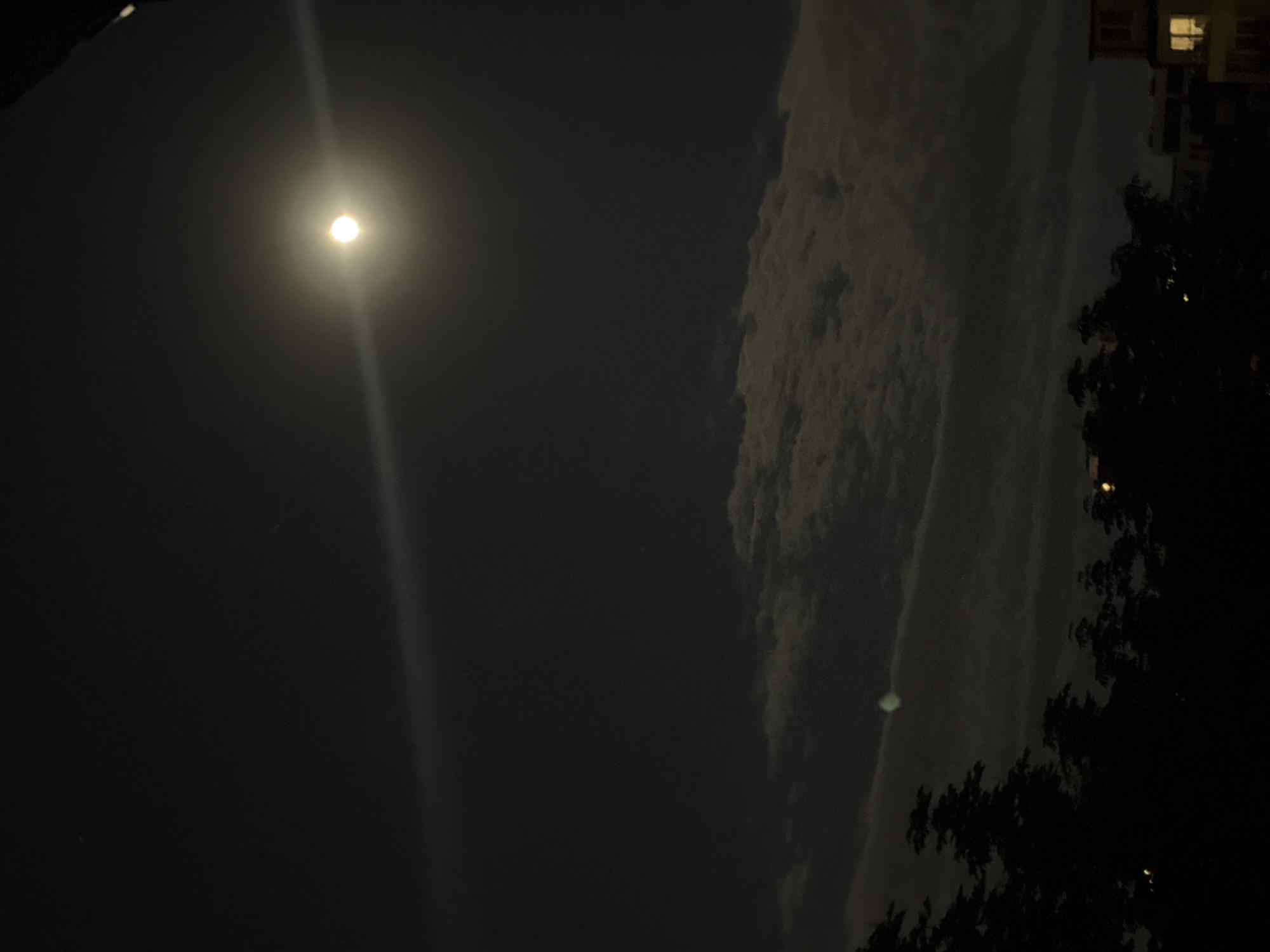 The image captures a night scene, likely in Sofia, Bulgaria. A bright moon dominates the upper left portion of the frame, with a prominent light ray extending downwards. To the right, a large mass of clouds is visible, partially illuminated by an unseen light source. A silhouette of trees occupies the far right, and a building with lit windows is partially visible at the top right corner. The overall atmosphere is dark and suggestive of an urban environment at night.