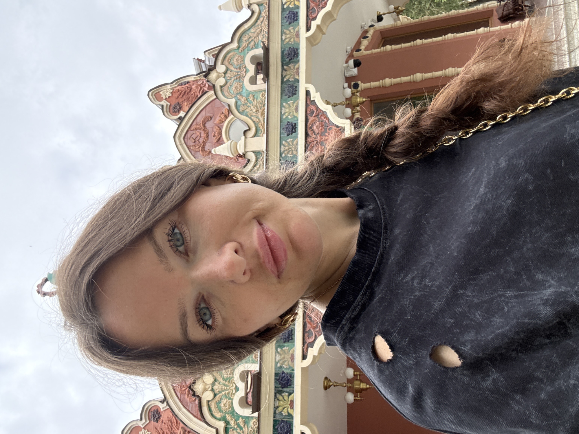 A close-up selfie is captured from a low angle, showing a woman with light brown hair, styled in a braid that drapes over her shoulder. Her eyes, a striking blue, are looking directly at the camera. Her lips are slightly parted, and she has a subtle smile. She is wearing a dark, possibly black, distressed t-shirt with two large circular cutouts on the torso. A gold chain strap is visible on her shoulder.
Behind her, an ornate building facade is visible, characterized by intricate decorative elements in shades of red, green, blue, and gold. The architecture suggests a grand or historical structure, possibly a palace or a museum, with arched doorways and detailed carvings. The lighting suggests it is daytime, but the sky in the upper portion of the image is overcast. The overall impression is one of a casual yet visually interesting setting, with the woman's modern attire contrasting with the detailed historical backdrop. No text is visible in the image. The location context is provided as Moscow, Russia.