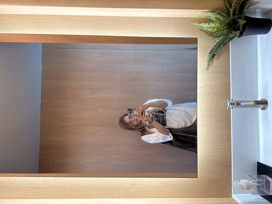 A person is reflected in a large, rectangular mirror with a wooden frame and soft, built-in lighting. The person is holding a smartphone and taking a picture of themself. The reflection shows a white t-shirt, glasses, and long brown hair. To the right of the mirror is a white sink with a metal faucet and a green fern in a black pot sitting on the countertop. Below, partially visible, is a soap dispenser. The background appears to be a wood-paneled wall, also seen in the mirror's reflection.
