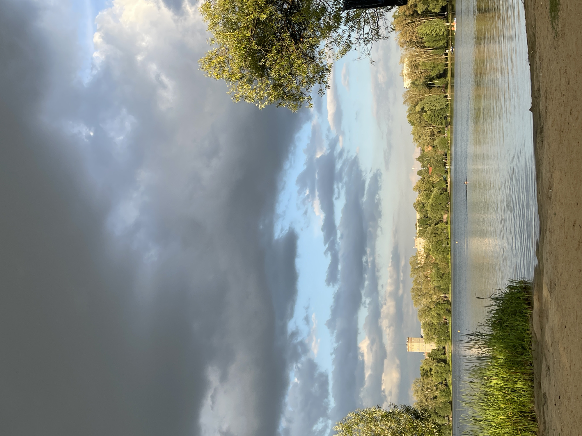 A body of water is shown with a sandy shore in the foreground. Lush green vegetation grows along the water's edge. A dense line of trees forms the background, with buildings and a tower visible behind them. The sky is filled with dramatic clouds, with patches of blue sky showing through. A person is swimming in the water, and two paddle boats are visible further out.