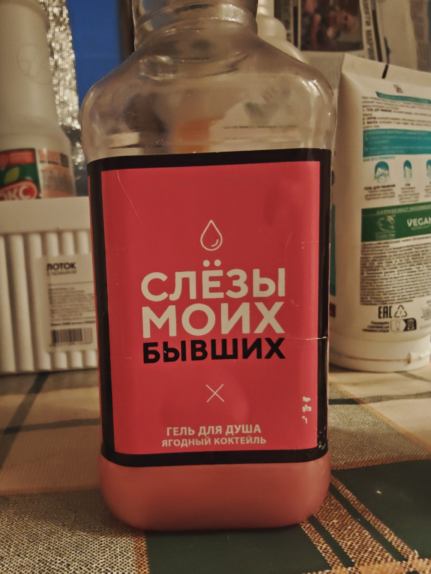 A close-up, eye-level shot shows a clear plastic bottle with a red label, filled with a pink liquid. The bottle is centered in the frame and rests on a checkered tablecloth. The label is predominantly red with white text and a small droplet icon at the top. The text on the label reads, in Russian, "СЛЁЗЫ МОИХ БЫВШИХ," which translates to "TEARS OF MY EXES." Below this, in smaller text, it says "ГЕЛЬ ДЛЯ ДУША ЯГОДНЫЙ КОКТЕЙЛЬ," meaning "SHOWER GEL BERRY COCKTAIL." To the right of the bottle, a white tube with a green stripe and text is partially visible. To the left, a white object with vertical ridges and a small label is also visible. The background is blurred but suggests a domestic setting. The lighting is dim, casting shadows and highlighting the texture of the tablecloth.