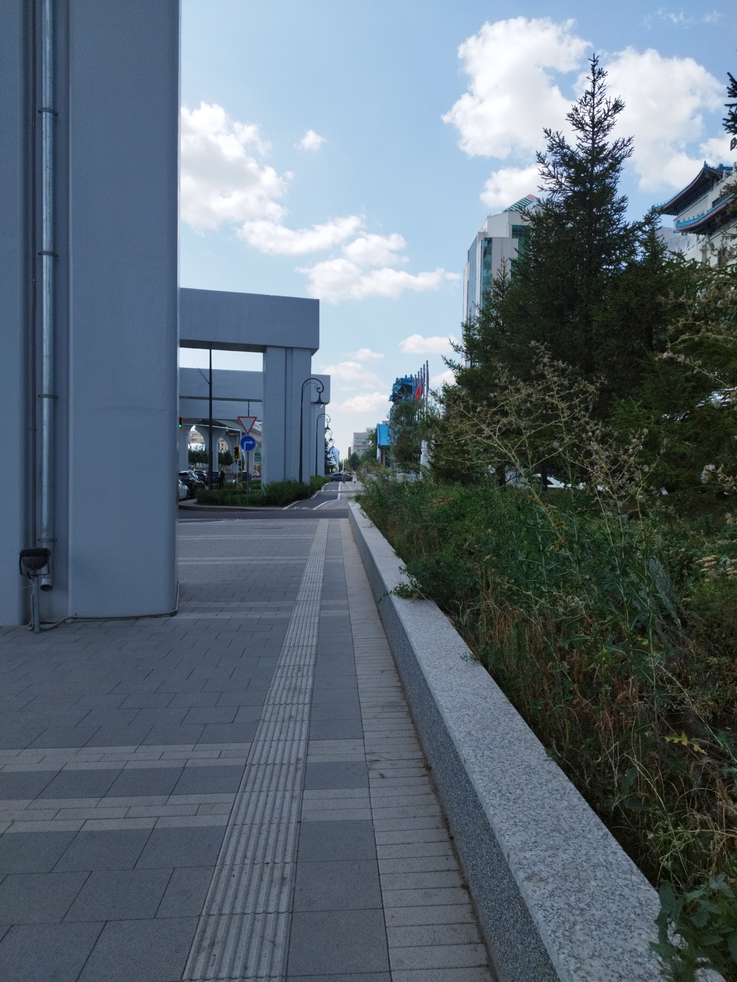 The image depicts a city street scene in Astana, Kazakhstan. In the foreground, a paved walkway with textured tactile paving runs alongside a concrete planter with a granite coping. To the left, a large, light-colored building with a metal conduit pipe running vertically up its facade dominates the frame. Beyond the walkway, a street with a traffic light and various cars is visible. Further in the distance, a tall evergreen tree stands prominently, with modern buildings, including one with a distinctive green roof, and traditional-looking architecture with blue tiled roofs also visible. The sky is blue with scattered white clouds, indicating daytime. The scene is a typical urban environment with infrastructure and greenery.
