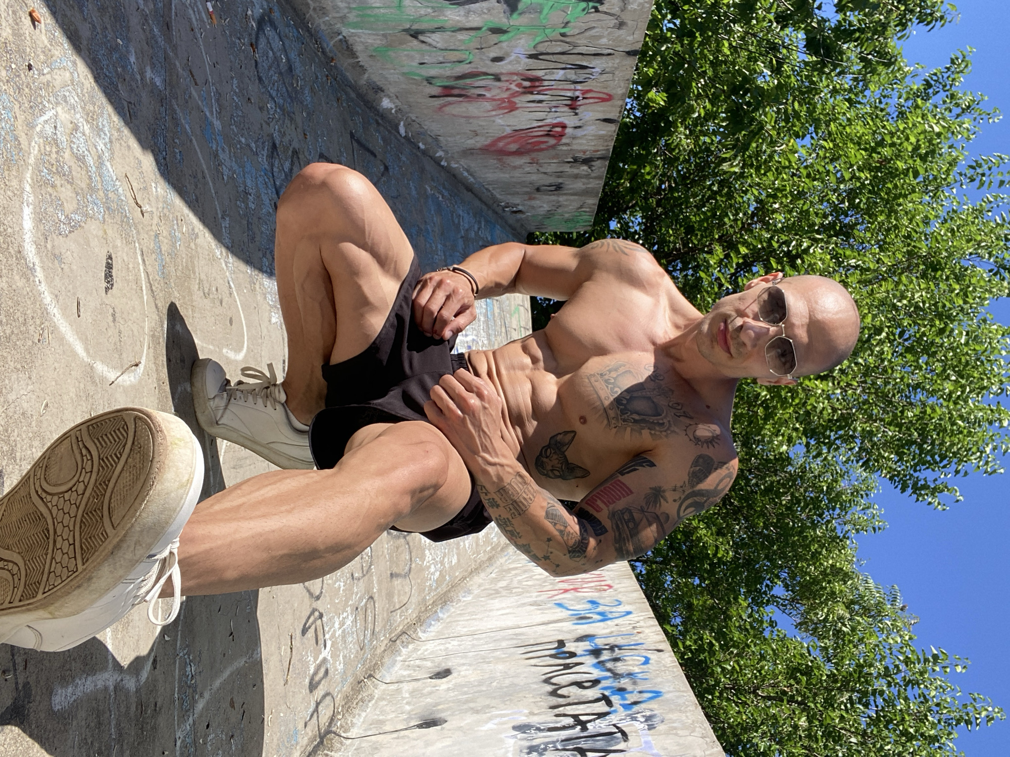 A bald, muscular man with tattoos on his chest and arms squats in a narrow outdoor walkway. He wears sunglasses and black shorts, and his white sneakers are visible. The walkway has a concrete floor with graffiti on the walls lining it. Lush green trees and a clear blue sky are visible in the background. The location is the former mausoleum of the Pleven Renaissance in Pleven, Bulgaria.
