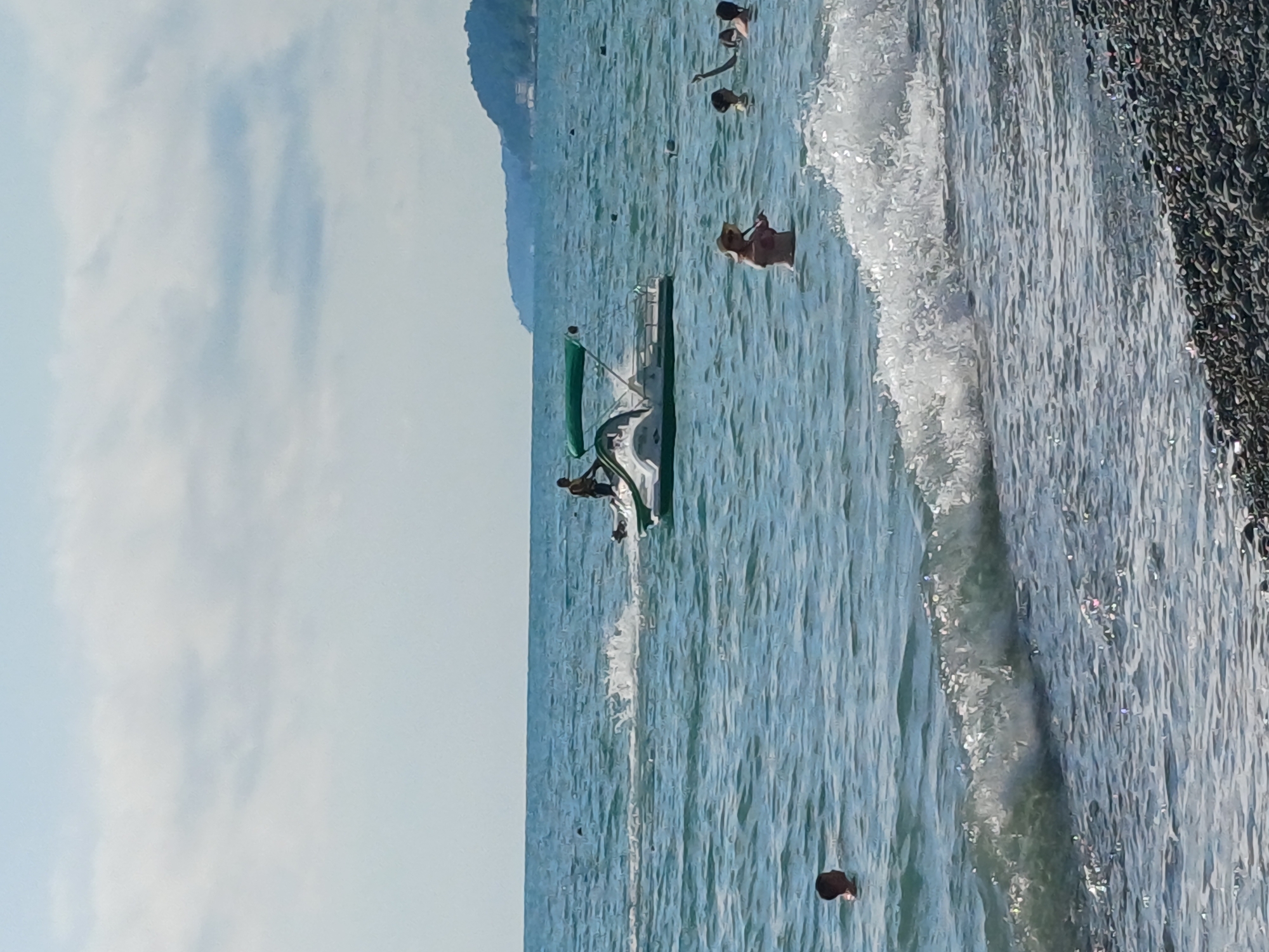 This media file captures a summer day at a beach in Sochi, Russia. The scene is dominated by the sparkling turquoise water of the Black Sea, with gentle waves rolling towards a pebbled shore. The sky above is a pale blue, with wispy white clouds hinting at fair weather.
Several people are enjoying the water and the beach. In the shallow water, a person can be seen standing and holding a straw hat. Further out, a small, white boat with a green canopy is visible. Two individuals appear to be near the boat, one possibly climbing out or in. Several other swimmers are scattered throughout the water. On the far right of the frame, the dark, glistening pebbles of the beach are clearly visible, with a gentle wave breaking against them.
The overall atmosphere is one of leisure and relaxation on a warm, sunny day. The light suggests it is daytime, likely late morning or early afternoon. There is no visible text in the media file.
