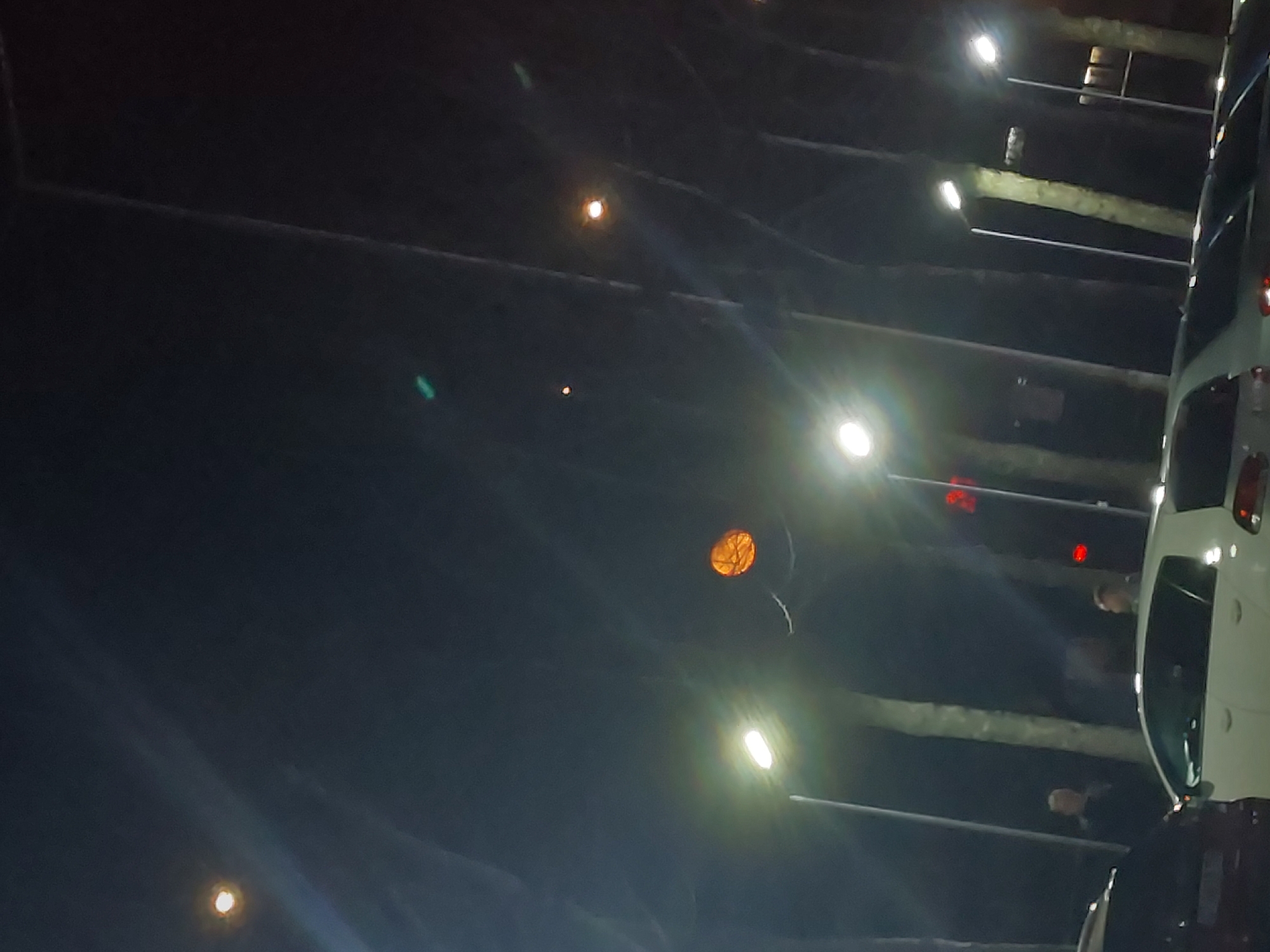The image, possibly taken at night in Asenovgrad, Bulgaria, shows a dimly lit outdoor environment with several light sources. An orange spherical object, possibly a balloon or decorative lantern, is suspended in the air. A row of vehicles, primarily white, are parked along the right edge of the image, illuminated by the surrounding lights. Indistinct human figures are visible near the vehicles. The background appears dark, suggesting a night scene or a poorly lit area. Horizontal beams or structures are discernible above the vehicles, possibly part of a parking structure or architectural feature.