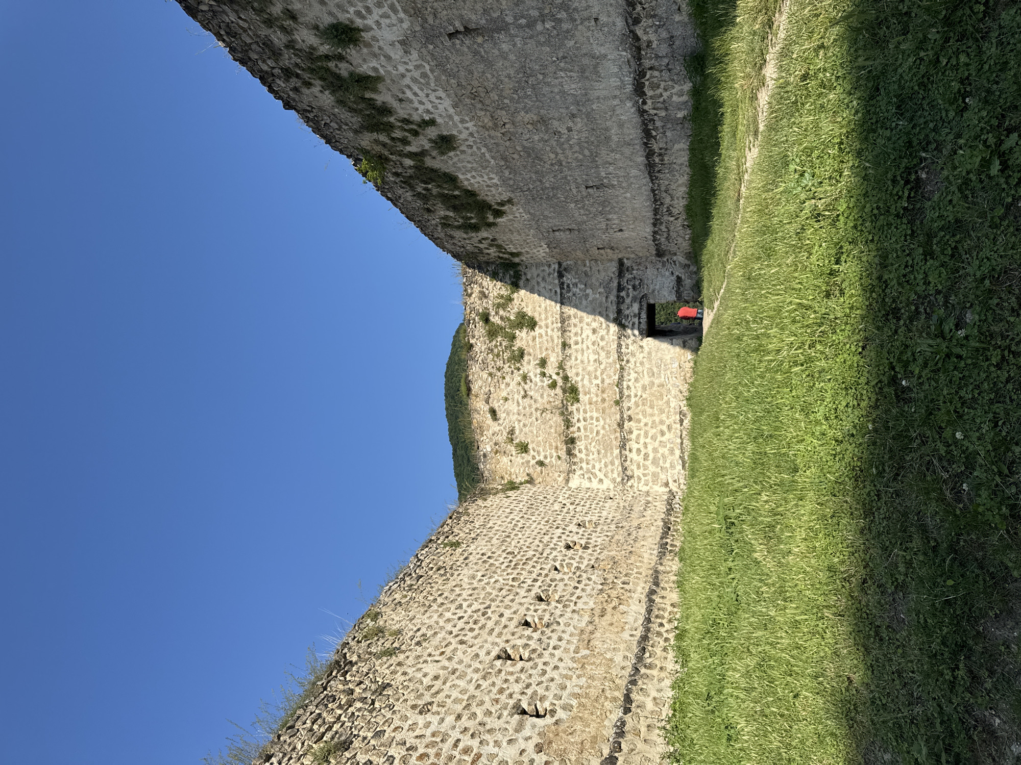 The image shows a view of the Khuchni fortress in Russia under a clear blue sky. The fortress walls are made of stone and are overgrown with vegetation in places. A person wearing a red jacket is seen standing in a doorway in the distance, within the fortress walls. The foreground is covered in green grass, with shadows cast by the walls.