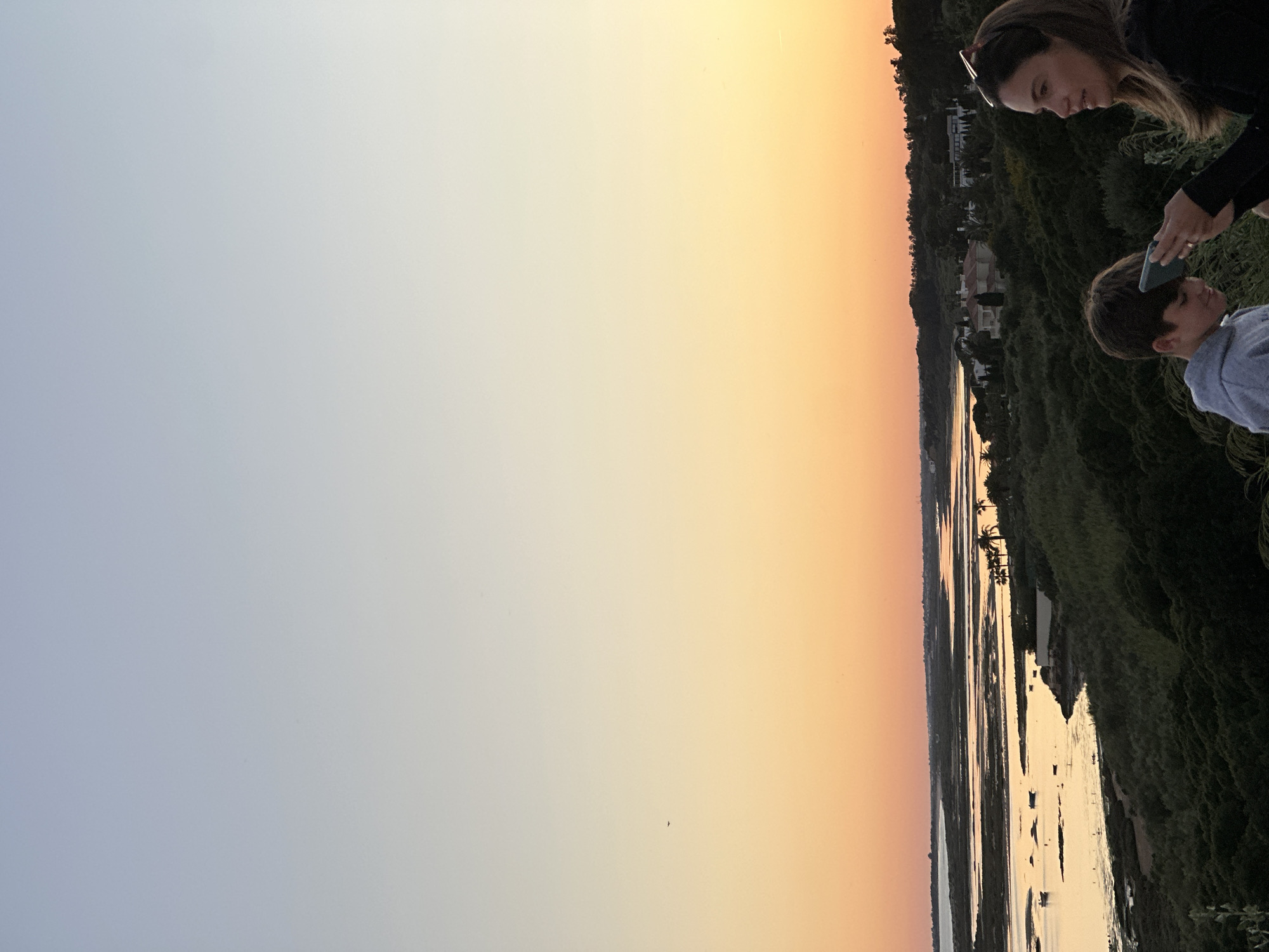 The image depicts a sunset view from a location overlooking a body of water in Vila Real de Santo António, Portugal. A woman and a boy are positioned on the right side of the frame, observing the scene. The woman is holding a phone near the boy's head, possibly taking a picture or showing him something on the screen. The backdrop features a blend of natural elements and man-made structures. There is a coastline densely covered with vegetation and houses, a mix of wetlands, channels, and water surfaces, and the sky during sunset is a gradient from light blue to orange.