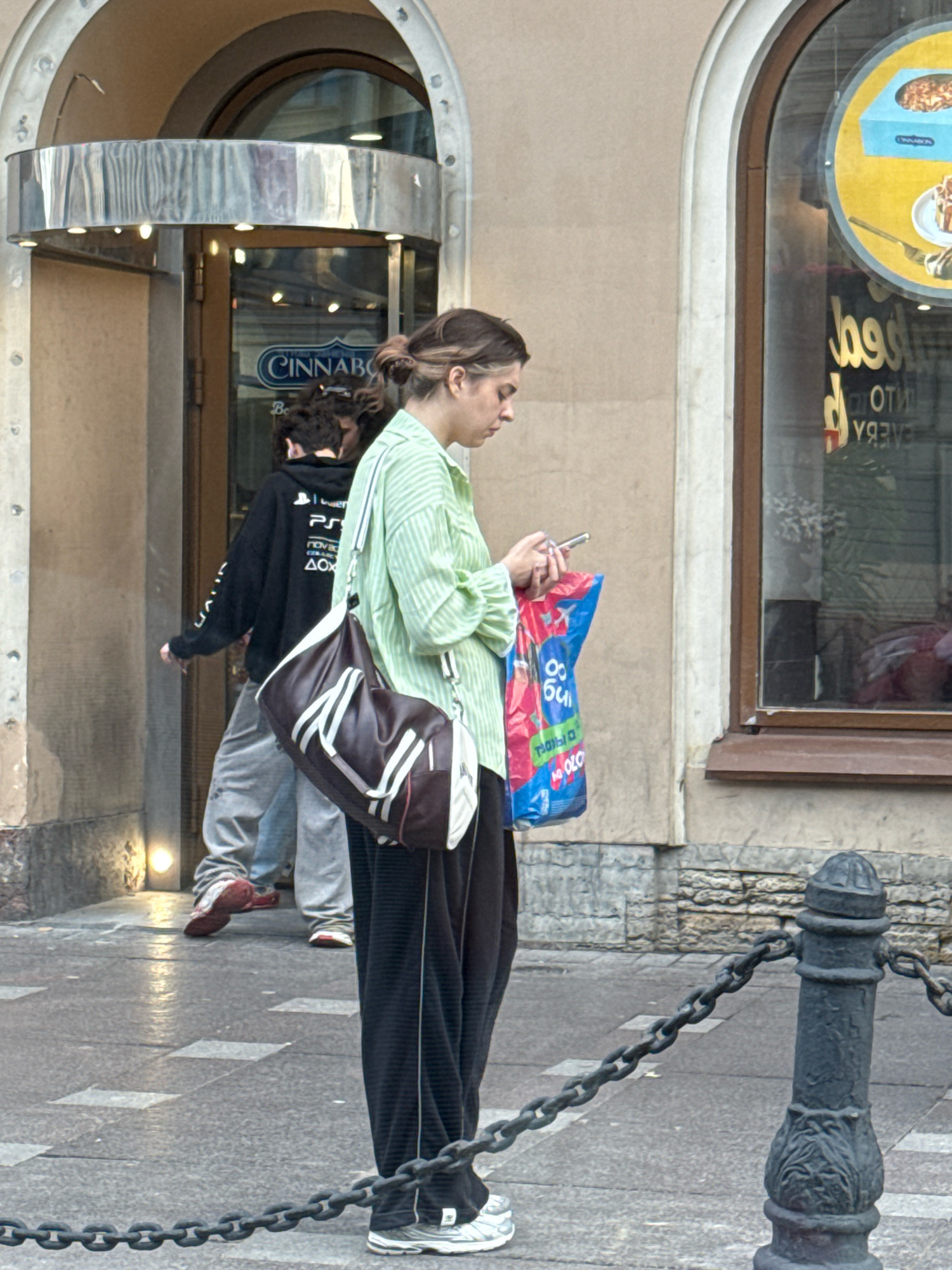 A woman in a light green striped shirt and black pants stands on a sidewalk in front of a building. She is holding a cell phone in her right hand and a red, blue, and white plastic shopping bag in her left. A dark brown and white duffel bag is slung over her left shoulder. Behind her, to the left, a person in a black hoodie and grey pants walks through a doorway. The doorway has a sign above it that reads "CINNABON". To the right of the woman is a storefront window with a yellow and blue circular advertisement. The sidewalk is paved with grey stones and has a metal chain barrier in the foreground. The architecture suggests the Nevsky Prospekt area of Saint Petersburg, Russia.