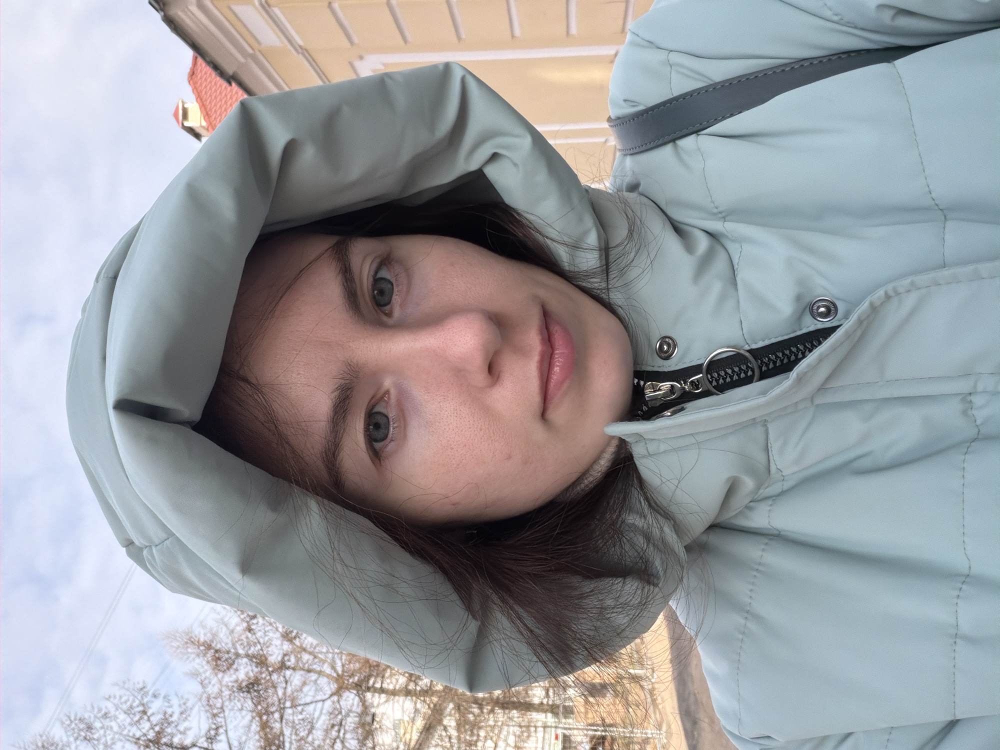 A woman with fair skin, light eyes, and dark hair is the main subject of the image. She is wearing a light blue, puffy winter coat with the hood up. The zipper of the coat is visible, along with snaps and a gray shoulder strap.
The background includes bare trees, a cloudy sky, and the top of a building with a red roof. The setting appears to be an urban outdoor location. It is difficult to determine the exact location in St. Petersburg without additional context, but the architectural style suggests an older part of the city.