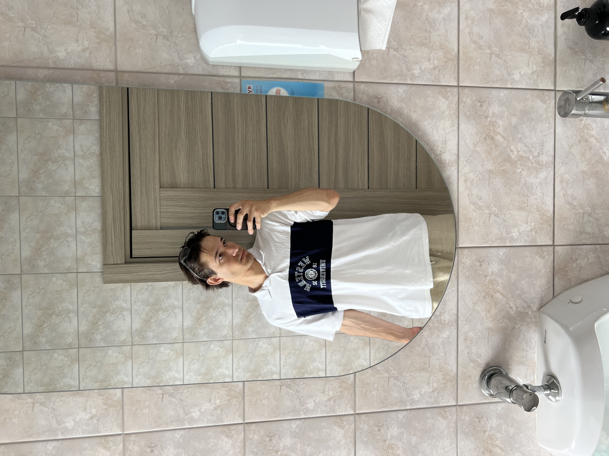 A young man is taking a selfie in a bathroom mirror. He is wearing a white polo shirt with a navy blue stripe across the chest that has "WESTERN UNIVERSITY" and other text in a circular logo. He has sunglasses perched on top of his head and is wearing light-colored shorts. The bathroom has tiled walls and a wooden door in the background. A white paper towel dispenser is mounted on the wall to the right, and a white urinal with a chrome faucet is visible in the lower left of the frame. The image appears to have been taken in a public restroom, possibly in or near Ждановіцкі сельскі Савет, Belarus.