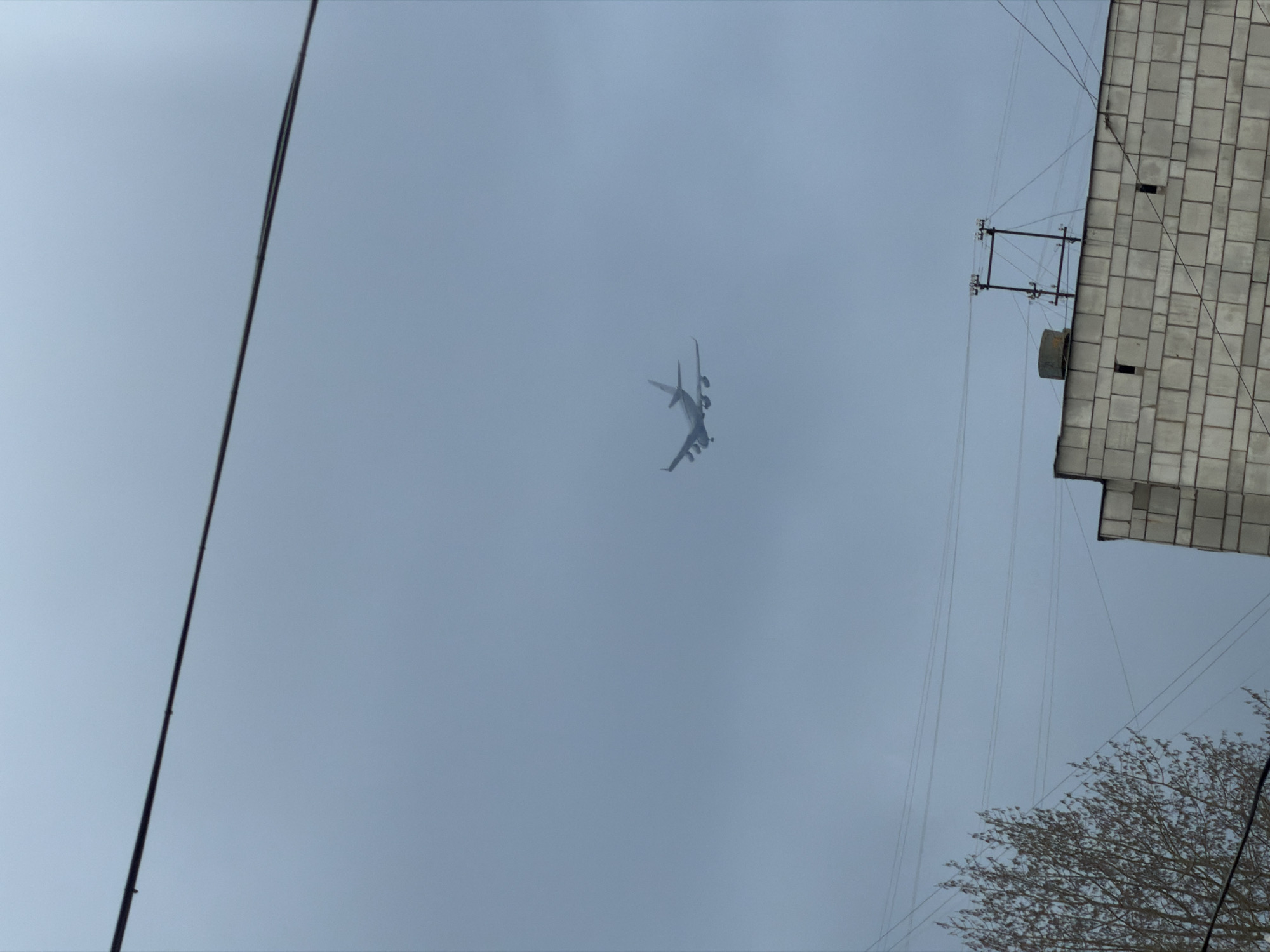 An outdoor, low-angle photograph captures a cloudy sky over what appears to be an urban or suburban environment in Perm, Russia. The image is tilted, presenting a light grey, overcast sky that dominates the frame.
A large, multi-engine aircraft, likely an airliner, is visible in the upper-middle left, flying across the sky. Its silhouette is somewhat indistinct against the muted background due to distance and atmospheric conditions.
In the foreground, a thick, dark utility cable or wire runs diagonally from the top to the bottom on the far left side of the image. On the right side, a section of a building is visible, featuring a facade constructed from grey, rectangular tiles or blocks. Numerous thinner wires and cables are strung across the sky near this building, some connected to a metal support structure protruding from the building's side. The top of a deciduous tree, largely bare of leaves, occupies the lower right corner, suggesting a non-summer season.
The time of day is daytime, indicated by the overall light, but the overcast conditions mean there are no strong shadows. No people or visible text are present in the image.