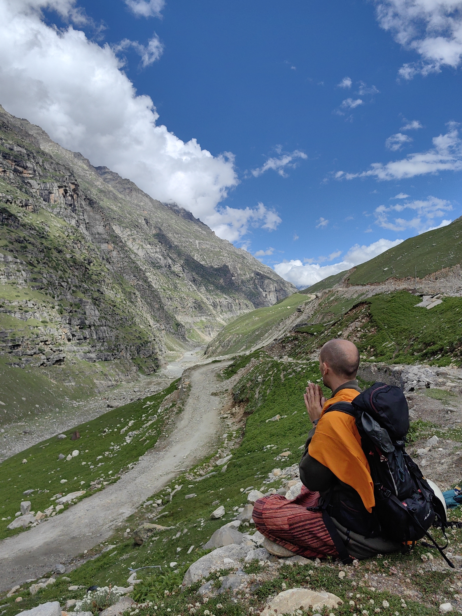 A bald man wearing an orange robe and striped pants sits with his hands pressed together in prayer. He is facing away from the camera, looking out at a mountain valley in India. The man has a large backpack on, and he is seated on rocks and grass. The valley is steep and green, with a dirt road winding through it. The sky is blue with white clouds.