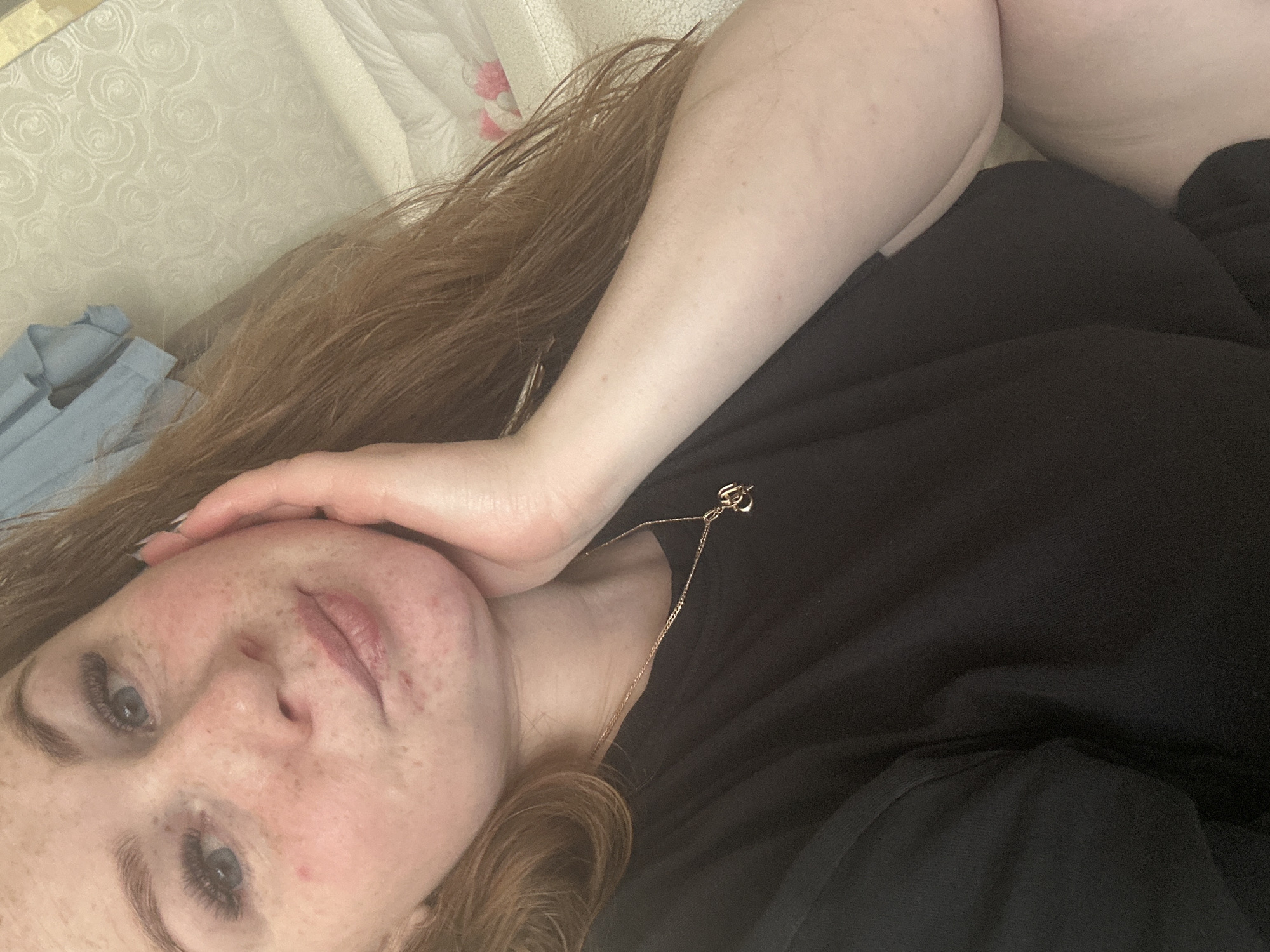 A selfie of a woman with freckles and red hair is shown. She is lying down, with her head resting on her hand. She is wearing a black shirt and a gold necklace. The background appears to be a textured, light-colored surface, possibly bedding or a wall. There is a blue garment visible in the lower-left corner. The location of Chaikovsky, Russia, is not evident in the image.