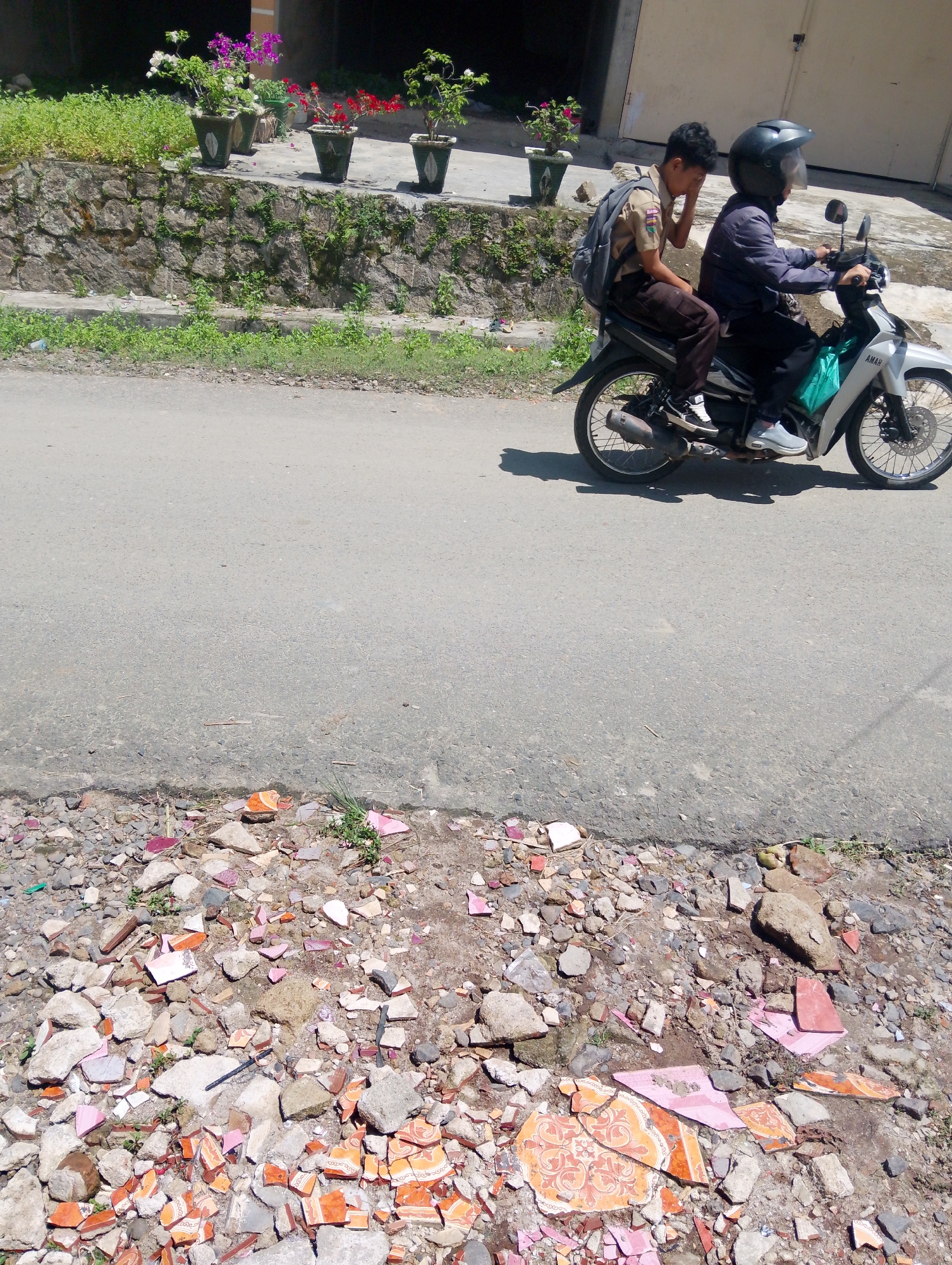 Two young Indonesian males are riding on a white motorcycle on a paved road in Serbelawan, Indonesia. The passenger, wearing a school uniform and backpack, appears to be resting his head against his hand. The driver is wearing a dark jacket and a black helmet. In the background, there is a stone wall with potted plants and lush green foliage. The foreground of the image shows a rough, debris-strewn area along the side of the road, with broken tiles and rocks scattered about.