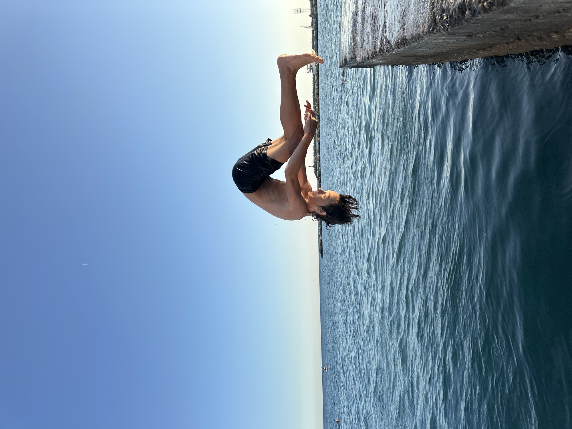 A young person is captured mid-flip, performing a dive from a concrete pier into the ocean. The individual is wearing black swim trunks and is oriented horizontally in the air, with their body arched and head pointing downwards. The ocean water is a deep blue with small waves, and the sky above is a clear, bright blue with a faint crescent moon visible. In the distance, a breakwater made of large stones extends into the sea, and beyond that, structures and a sailboat are faintly discernible. The scene appears to be an outdoor recreational area, possibly a pier or harbor, in Sochi, Russia, given the bright sunshine and coastal environment.