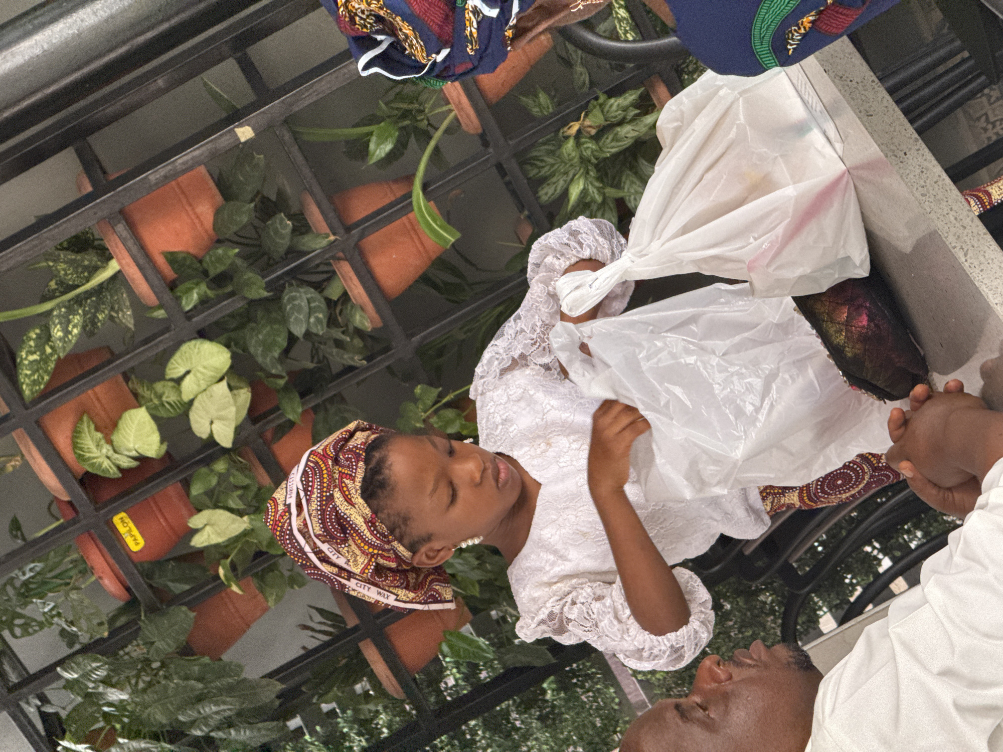 A young girl with a patterned headwrap and white lace dress is opening a white plastic bag outdoors. She is standing next to a man wearing a white shirt. They are near potted plants in a black metal structure. The headwrap is labeled "CITY WAX." The location is most likely in Abuja, Nigeria.