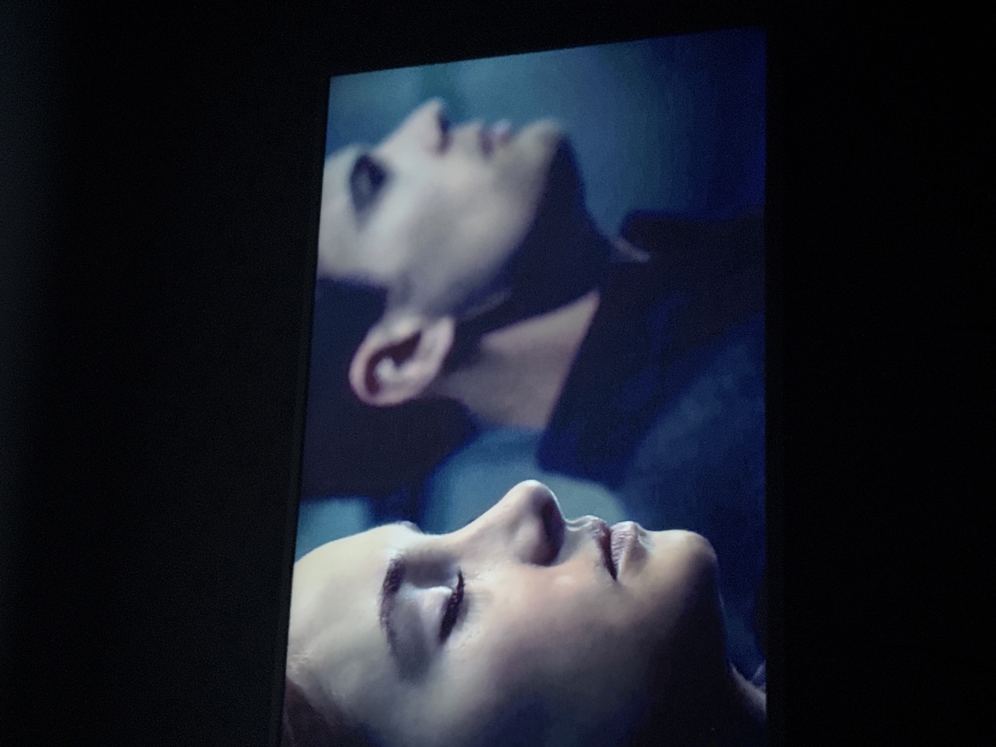 The image shows a close-up of two faces. The bottom face appears to be female, with closed eyes, facing upwards. Above and slightly behind is another face, possibly male, also looking upwards. The overall tone is blueish. No location in Mytishchi, Russia, is discernible.
