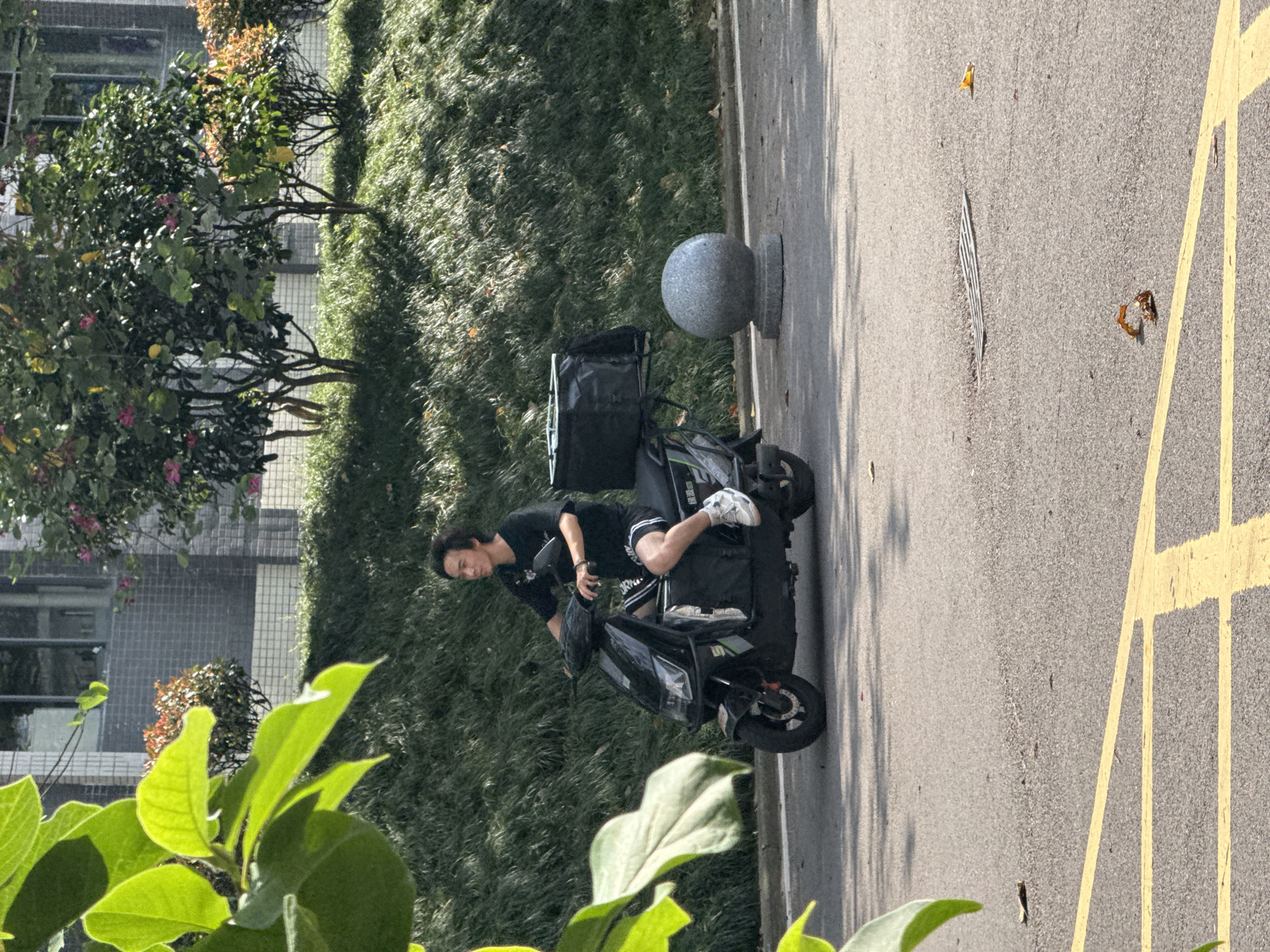 In Jiangjin, China, a young man is seated on a black electric scooter, parked on the side of a paved road. He is wearing a black t-shirt and shorts, with white athletic shoes. The scooter has two large black storage boxes mounted on the back. The scene is set with a lush green lawn bordering the road, and beyond the lawn, a building with a checkered facade. Trees with pink flowers are visible near the building. A grey spherical ornament on a pedestal stands near the scooter. The sunlight casts shadows, suggesting it is daytime. The overall mood appears casual and ordinary. There is no visible text in the image.