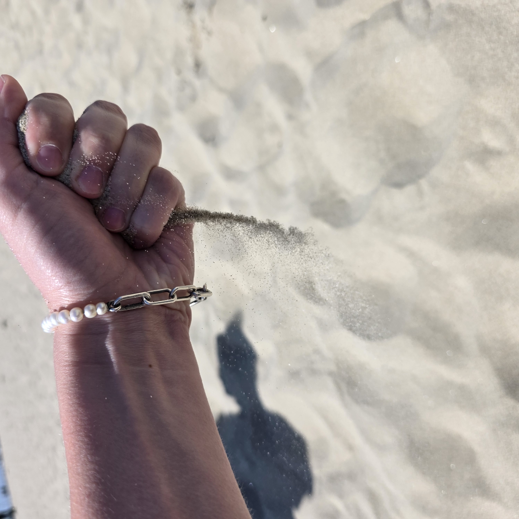 A close-up, low-angle shot focuses on a human hand cupped around sand, allowing it to trickle through the fingers onto a light-colored sand beach. The individual's hand is adorned with a bracelet featuring pearls and a silver chain. The background is a blurry expanse of sand, suggesting a beach environment. The scene is set in Cannon Beach, United States of America. The lighting indicates it is daytime.