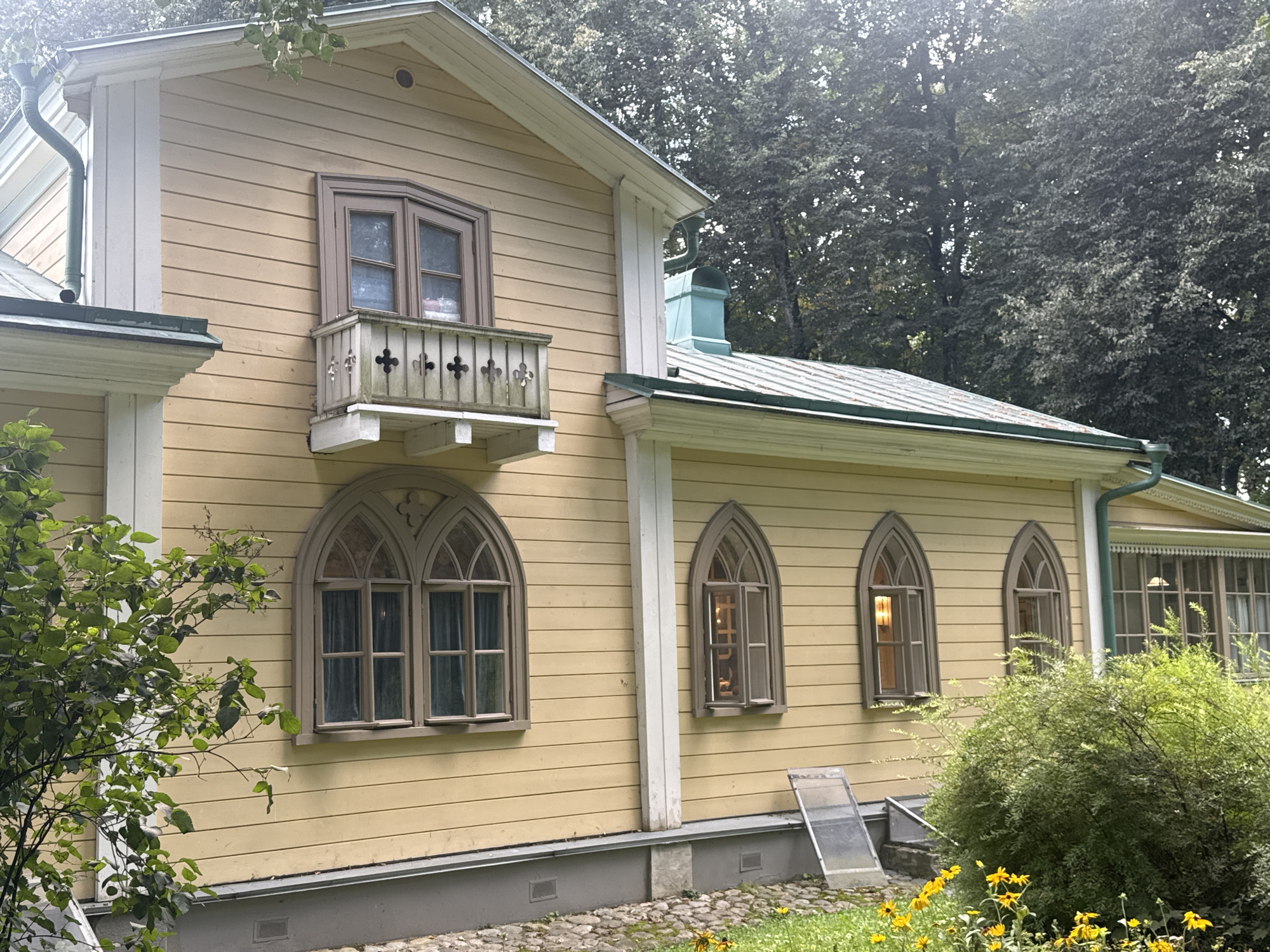 The image depicts the exterior of a light yellow wooden house located in the village of dacha cooperative "Arkhangelskoye," Russia. The house features a light green gutter system and a metal roof. Prominently visible are several arched windows with dark brown frames. One of these windows is a bay window on the upper floor, adorned with a small white balcony featuring decorative wooden cross shapes. On the lower level, there are three Gothic-style arched windows, two of which are large and joined by a narrow white column, and a single narrower arched window to their right. One of the lower arched windows is open, revealing interior lighting. To the right of this window, a large green bush with dense foliage partially obscures the lower part of the house. In the foreground, yellow wildflowers are scattered amongst green grass and a cobblestone pathway. Two clear plastic sheeting pieces are leaning against the house near the lower windows. The background is dominated by a dense canopy of tall, dark green trees. The lighting suggests it is daytime, with some sunlight filtering through the trees, creating a slightly hazy effect.