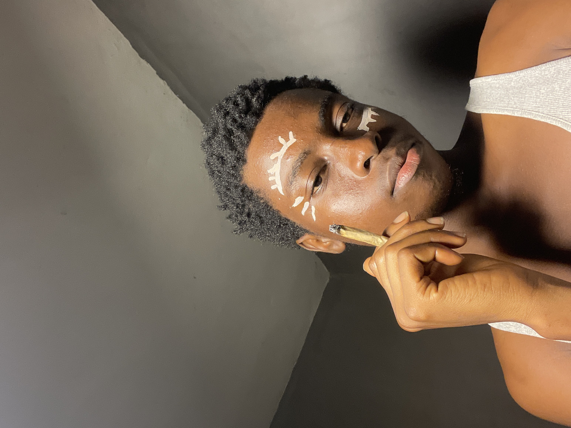 In Nkpor, Nigeria, a young Black individual with dark, curly hair is shown in a close-up, low-angle shot. They are wearing a white ribbed tank top and have white paint applied to their face in an artistic design around their eyes and forehead. The individual is holding a lit cigarette with their right hand, bringing it towards their mouth. The background consists of a dark gray wall and a lighter gray ceiling.