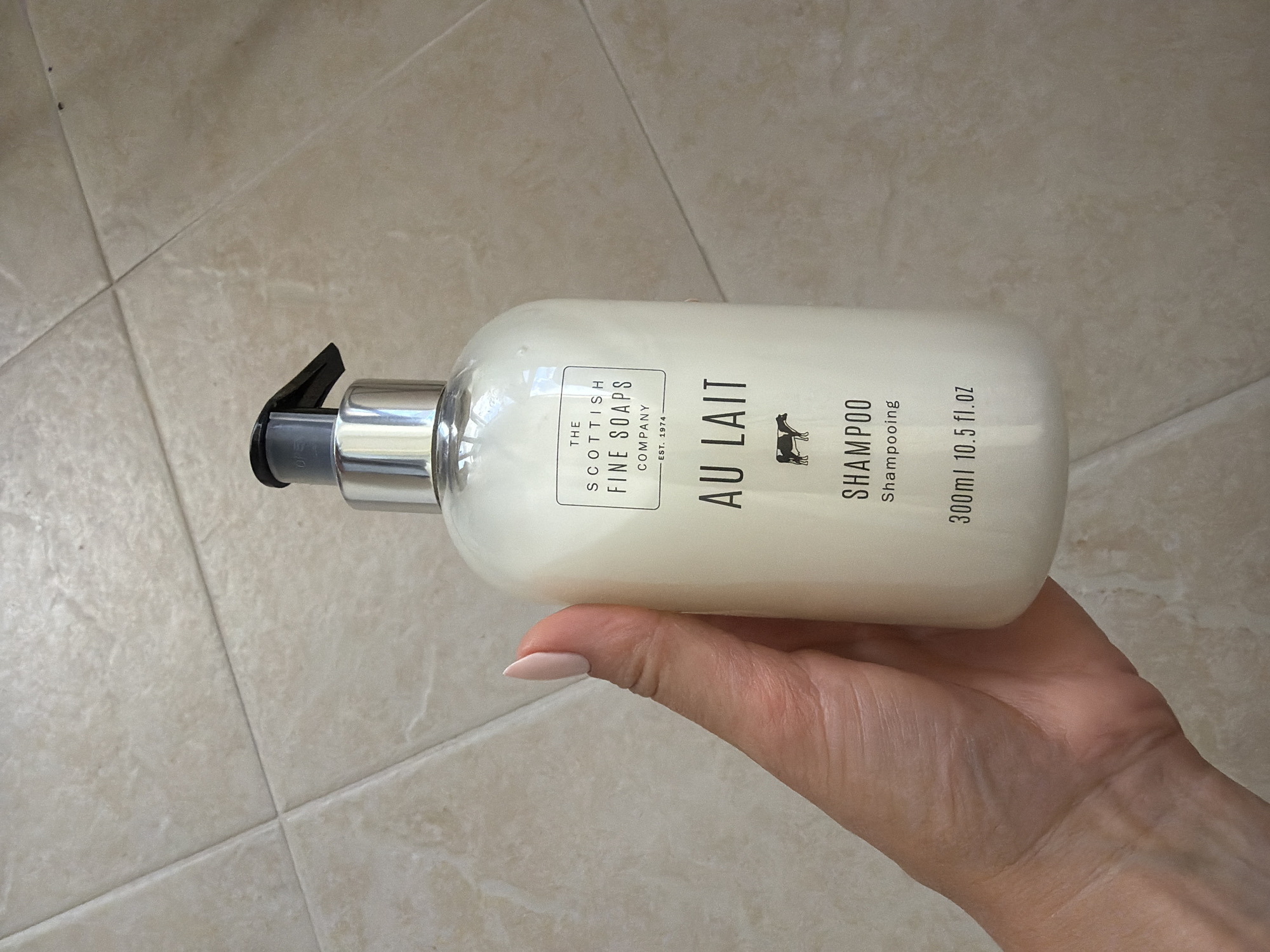 A person is holding a white bottle of "Au Lait Shampoo" by The Scottish Fine Soaps Company. The bottle has a black pump dispenser and a label that includes the brand name, product name, and volume (300ml / 10.5 fl.oz). The background appears to be a tiled surface. The image was taken in Pazardzhik, Bulgaria.