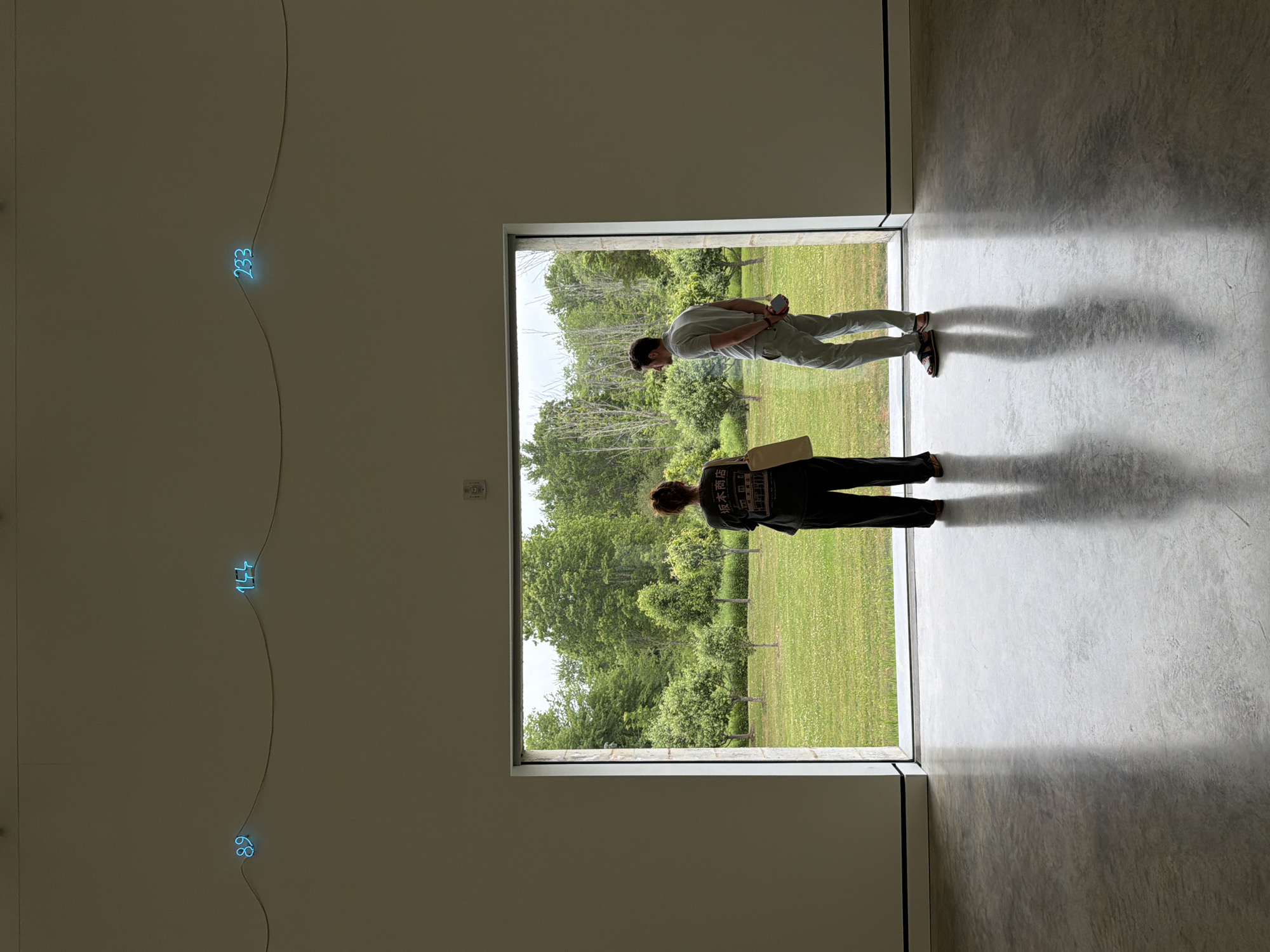 Two individuals, a woman and a man, stand silhouetted against a large rectangular opening in a white-walled interior, looking out into a sunlit garden. The woman, on the left, wears dark, loose-fitting pants and a graphic t-shirt, with her hair pulled back. She holds a light-colored bag. The man, on the right, is dressed in light-colored pants and a short-sleeved polo shirt. He faces away from the camera, looking out at the garden.
The interior features a polished concrete floor with prominent shadows cast by the two figures. Above the opening, three blue neon numbers are suspended from thin wires against the white wall: "89", "144", and "233".
The exterior view reveals a lush green lawn dotted with numerous small trees, suggestive of an orchard or manicured garden. Beyond the garden, a dense line of mature trees forms a natural backdrop under an overcast sky.
The image appears to be taken within a gallery or museum space. The specific location in Cold Spring, United States, is likely the Putnam County Visitors Bureau or a similar art exhibition space, given the contemporary art installation of neon numbers and the gallery-like setting.