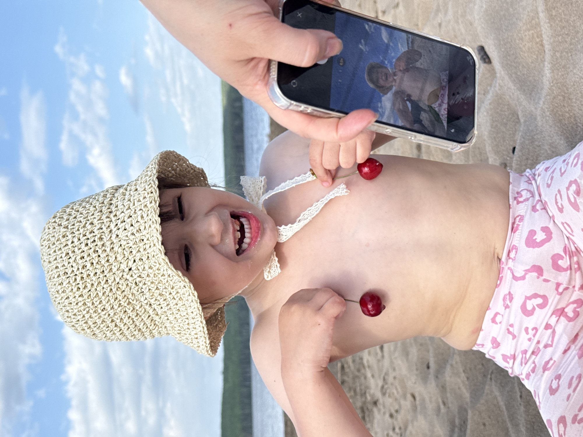 A rotated eye-level shot captures a young child on a sandy beach in what appears to be Chaikovsky, Russia. The child is wearing a straw hat and a white lace-trimmed top, with pink patterned shorts. The child is holding cherries on their chest, posing for a photo being taken with a smartphone. The phone's screen shows a live image of the same scene, creating a picture-in-picture effect. The background includes a body of water, trees, and a blue sky with clouds, indicating an outdoor, possibly vacation-related setting.