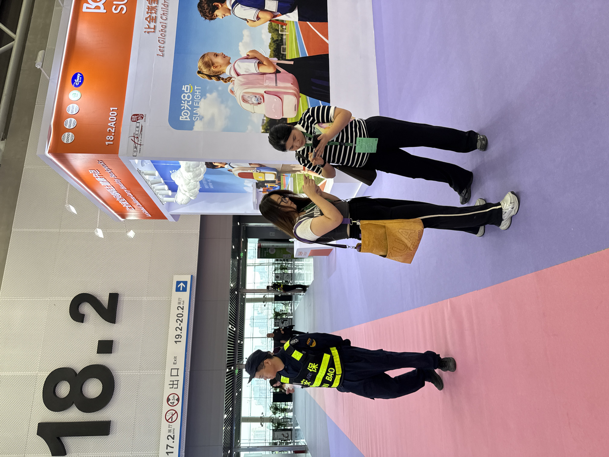 This is a photograph taken at an exhibition hall, likely in the Haizhu District of China, judging by the presence of signage in Chinese and English. The scene is brightly lit and appears to be a trade show floor with a large, colorful display booth in the background.
In the foreground, a security guard wearing a dark uniform and a bright yellow vest with Chinese characters and "IN BAO" written on it is standing on a pink carpeted pathway. They are looking to their left with a neutral expression. To the right of the security guard, two women are standing on a purple carpet. The woman on the left is wearing glasses and a dark outfit with a purple strap across her chest. She is looking down at her phone. The woman on the right is wearing a black and white striped shirt and dark pants, and she is also looking down at her phone, which is held in her right hand. She has a green visitor badge clipped to her shirt.
In the background, a large, orange and white booth is visible, advertising a product with the brand name "SUN EIGHT" and English text that reads "Let Global Children Carry Healthy Backpacks." There are several certifications displayed on the booth, including ISO 9001, ISO 14001, and a Disney logo. Images of children with backpacks are also displayed on the booth. Further in the background, more exhibition booths and a large open space with many potted plants are visible, suggesting a spacious venue. The overall environment is clean and organized, indicative of a professional exhibition setting.