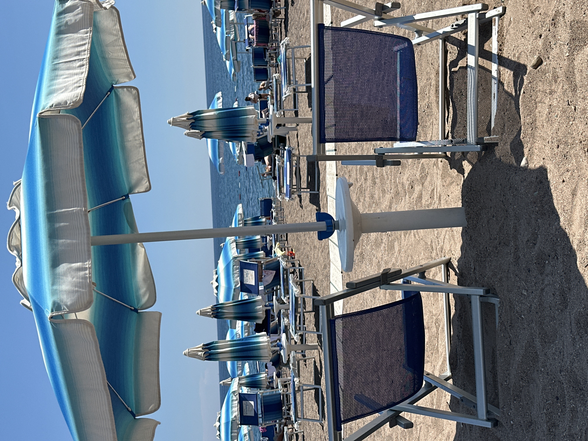 The image depicts a beach scene in Boreti, Montenegro, featuring rows of identical beach chairs with blue woven seats, arranged in pairs under matching blue and white striped beach umbrellas. The foreground shows two unoccupied chairs, casting distinct shadows on the sand. Further into the scene, more chairs and umbrellas stretch towards the coastline, where a sliver of sea is visible under a clear blue sky. Some people are visible in the distance amongst the chairs.