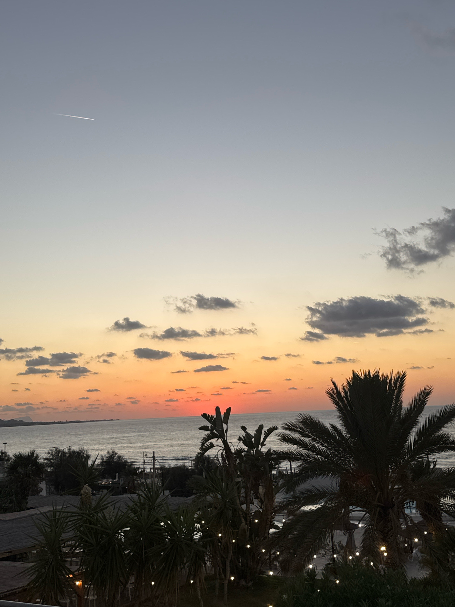 A serene sunset casts a warm glow over the sea, with the sun just dipping below the horizon, painting the sky in shades of orange and pink. The ocean is calm, reflecting the colors of the sky. In the foreground, lush palm trees and other tropical foliage create a silhouette against the vibrant sky. Scattered lights, possibly from an outdoor dining area or pathway, twinkle amongst the plants, adding a touch of ambiance. In the distance, a faint outline of land is visible, hinting at the coastline of Isola delle Femmine, Italy. A thin contrail from an airplane streaks across the pale blue upper sky.