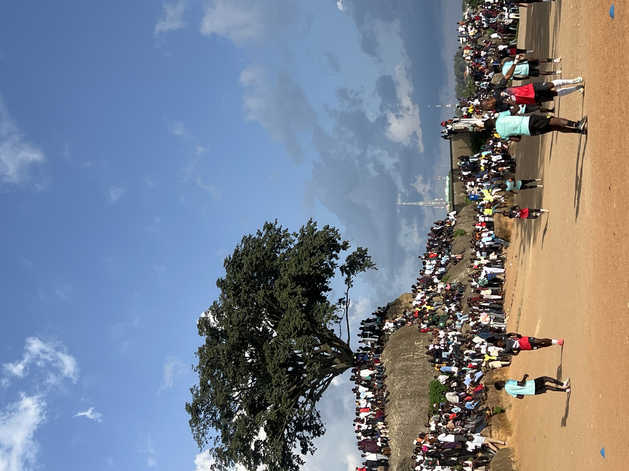 In Jos, Nigeria, a large crowd of spectators is gathered on a rocky, elevated terrain to watch a soccer match. The spectators are densely packed, with some sitting and others standing, creating a vibrant and engaged audience. The soccer field is a dirt surface, and several players are visible in action. A prominent, large tree with dense foliage stands on the rock formation, casting a shadow over part of the scene. The sky above is a bright blue with scattered white and gray clouds, suggesting it is daytime. The overall atmosphere appears to be one of a lively community event.