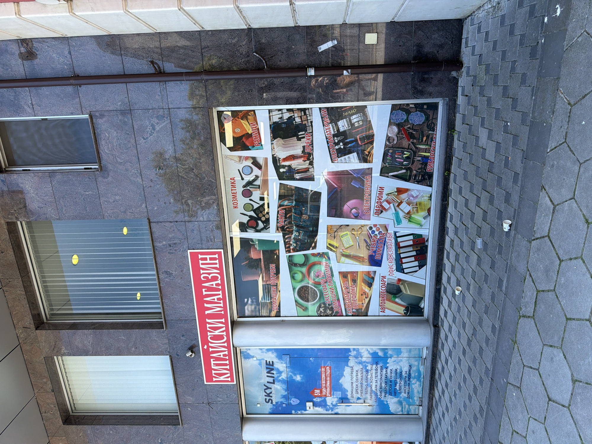 Here is a description of the image:
The image shows the exterior of a store located in Plovdiv, Bulgaria. The store's name is "Chinese Store" ("Китайски Магазин" in Bulgarian). The entrance is labeled "SKY LINE магазин" with a picture of clouds. A sign advertises a variety of goods available inside, including household items, bags, cosmetics, clothing, shoes, school supplies, underwear, swimwear, perfumes, accessories for phones, office supplies, fishing gear, toys, car accessories, electronics, and accessories for pets. The store's entrance is not directly accessible from this side of the building; a sign indicates that the entrance is on the other side of the building. The facade of the building is made of gray stone tiles. The store has windows with blinds. The sidewalk is paved with gray hexagonal and square tiles.