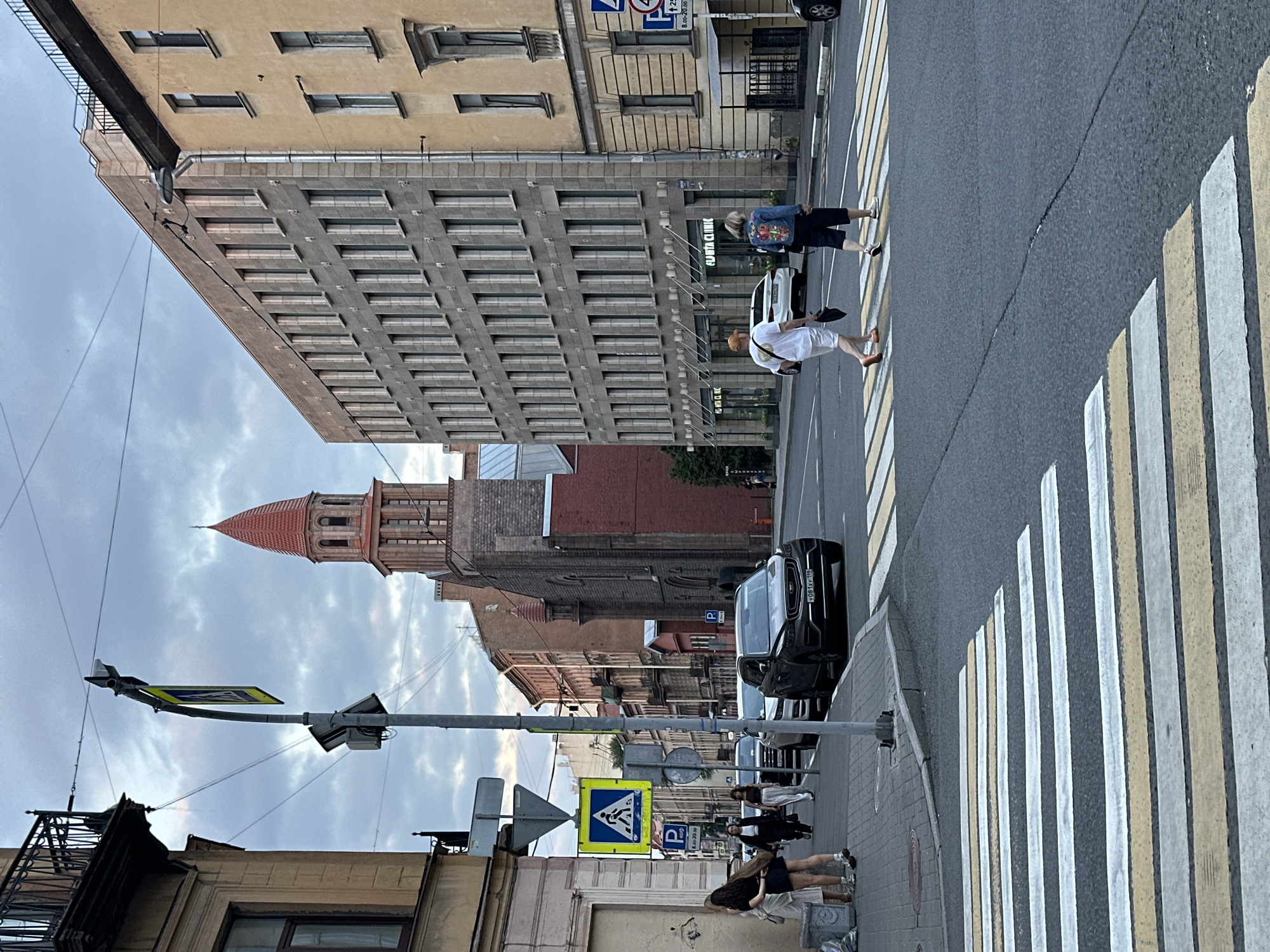 The image depicts a street scene in Saint Petersburg, Russia, likely during twilight. The perspective is from a street corner, looking down a road with a prominent pedestrian crossing in the foreground.
On the left side of the street, there are pedestrians waiting on the sidewalk, with a building and a traffic light pole with street signs visible. The traffic light pole displays a pedestrian crossing sign, indicating a designated area for people to cross the road. Further down the street on the left, a dark-colored SUV is parked.
Across the street, on the right side, a multi-story building with numerous windows dominates the scene. A narrower building with a red brick facade and a distinct red-tiled tower with a spire is visible behind it. This tower-like structure suggests a church or a historical building. Several people are crossing the street at the pedestrian crossing. One person in a white t-shirt and shorts walks with a bag, and another individual in a denim jacket and shorts walks alongside.
The road surface is asphalt, and the pedestrian crossing is marked with white and yellow stripes. The sky is overcast with clouds, characteristic of the Saint Petersburg climate. The overall atmosphere is that of a typical urban environment during the evening.