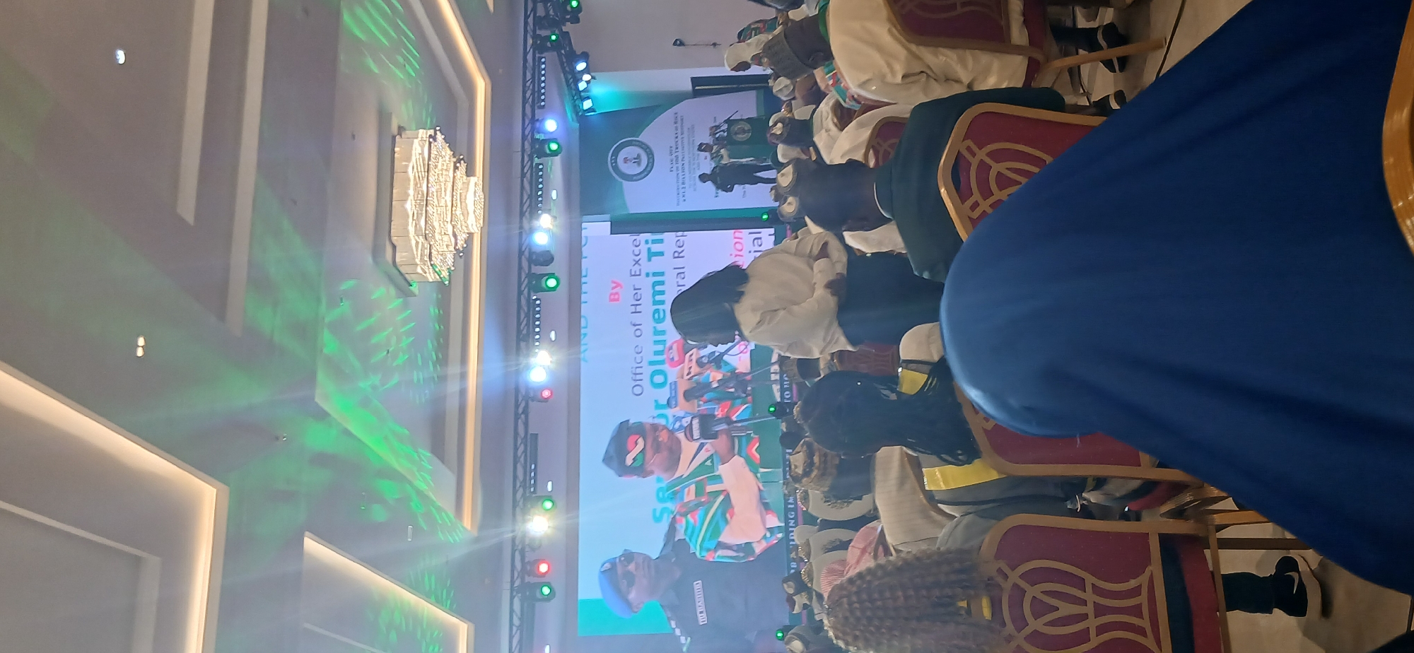 The image shows an indoor event in a large room, likely in Unguwan Sanusi, Nigeria, based on the onscreen text about Oluremi Tinubu. There is a large screen on stage displaying images and text. Several people are seated in chairs, facing the stage. A person stands at a podium on the screen, likely giving a presentation. The room has modern lighting, including a large chandelier, and green lighting effects on the ceiling.