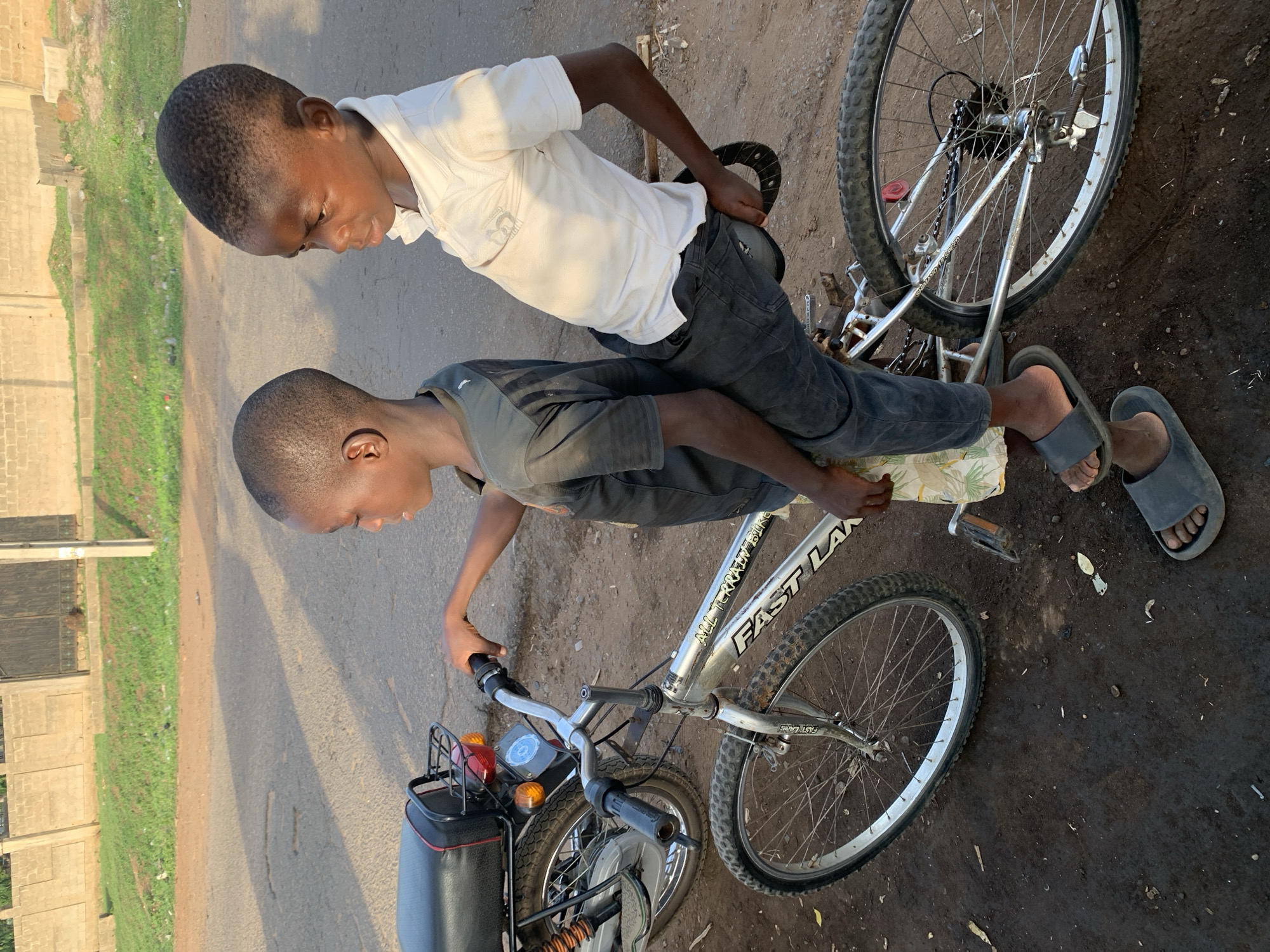 Two young boys are depicted near a bicycle in Kabba, Nigeria. One boy, wearing a white shirt and dark pants, sits on the seat of the bicycle. The second boy, wearing a dark shirt, stands by the handlebars. The bicycle is silver and has a carrier rack on the rear. The ground is dirt and pavement. In the background, there is a grassy area and a building.