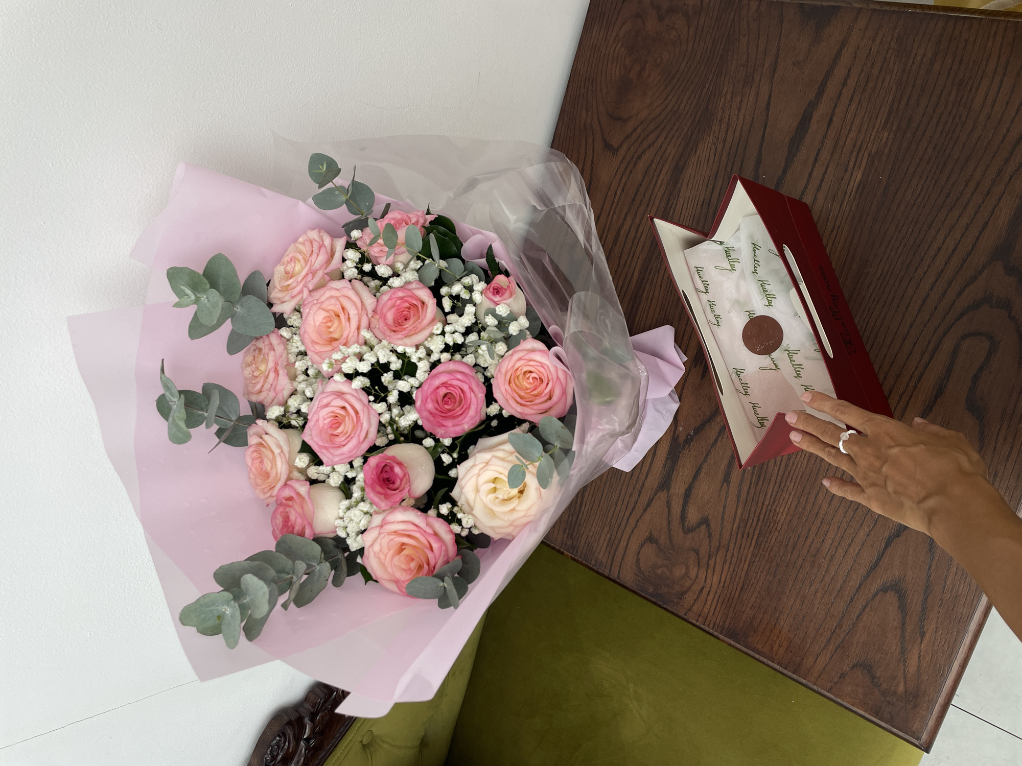 The image captures an indoor scene in Thủ Đức, Vietnam, featuring a bouquet of flowers, an open box, and a person's hand on a polished wooden table. The bouquet, centrally placed on the table, consists primarily of pink and light pink roses, interspersed with small white gypsophila (baby's breath) flowers and eucalyptus leaves. It is wrapped in layered pink paper and transparent plastic, with visible water droplets on the petals and wrapping. To the left of the bouquet, a portion of a green upholstered armchair with a dark, carved wooden armrest is visible. The background consists of a plain white wall.
In the lower right portion of the image, an open, rectangular red box with a white interior rests on the table. Inside the box, white tissue paper patterned with repeating cursive text, specifically "Healthy Healing," is visible, along with a circular brown sticker bearing a logo. A person's tanned right hand, adorned with a white multi-stone ring on the ring finger, extends from the bottom of the frame, reaching towards the red box. The dark wooden table exhibits prominent grain patterns. A small section of light-colored tiled flooring is discernible at the bottom left edge of the frame.