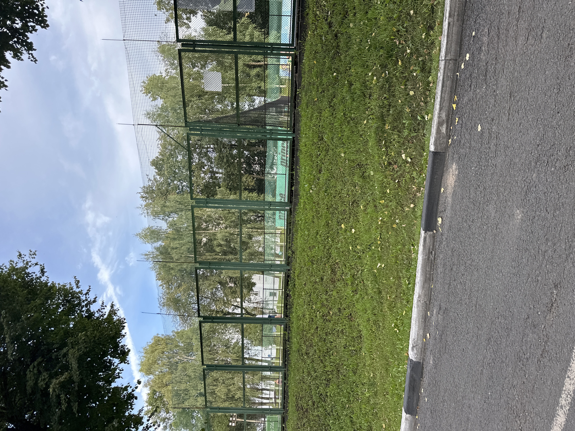 A paved road borders a grassy embankment topped by a green chain-link fence. Beyond the fence, a tennis court with blue and green surfaces is visible, along with a white building and trees. The sky is partly cloudy. The location is identified as Putyatino (Semivragovskaya S/A), Russia.