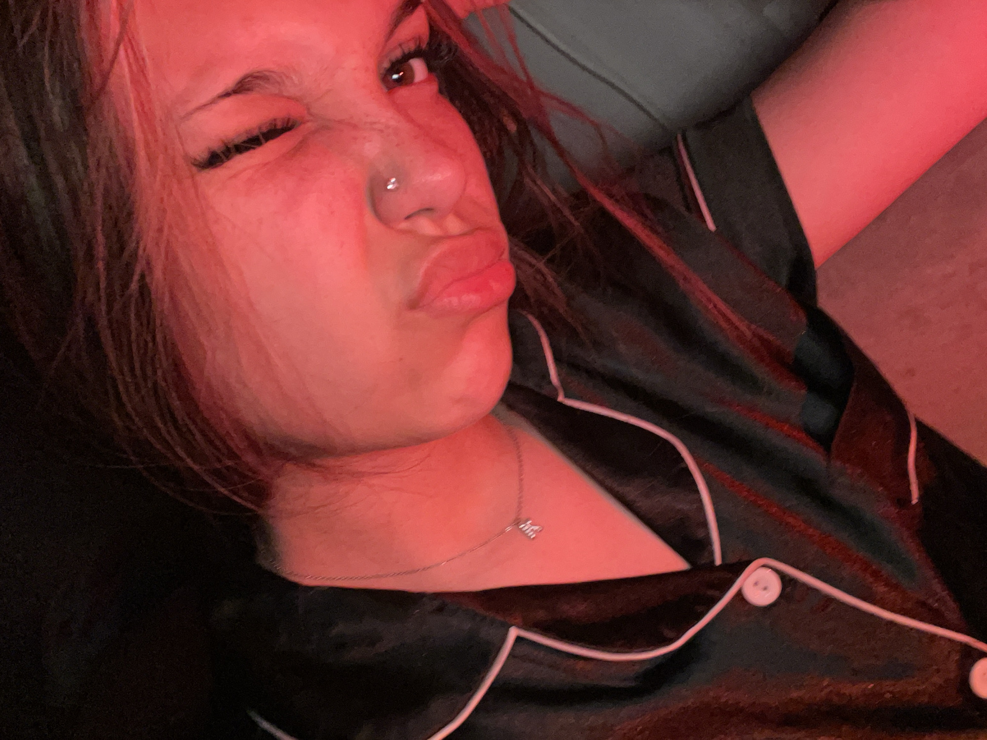 The image shows a close-up of a young woman's face illuminated by red light. She is wearing a black satin pajama top with white trim and buttons, and a delicate silver necklace with a small charm. She has a nose piercing and her eyes are partially closed, with her mouth slightly pursed. Her hair, which appears to be dark with some reddish highlights from the lighting, is falling around her face. The background is dark and indistinct. There is no information in the image to suggest a specific location in the city of Unknown, Russia.