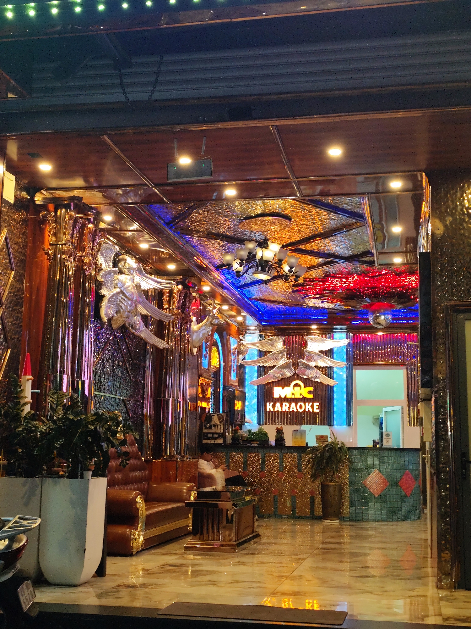 The image captures the entrance of the M.I.C. Karaoke establishment in Da Lat, Vietnam. The interior design is opulent, featuring mirrored and textured gold-colored walls, ceilings, and decorative columns. There are ornate angel sculptures on the walls, and a chandelier hangs from a textured ceiling illuminated with blue and red lighting, along with a disco ball. The reception area has a turquoise counter with diamond patterns and is lit by a blue LED sign displaying "M.I.C. KARAOKE." A brown leather couch and wooden coffee table are on the left. Houseplants in white pots and a motorcycle are visible in the foreground outside the entrance. A person is seated behind the reception counter.