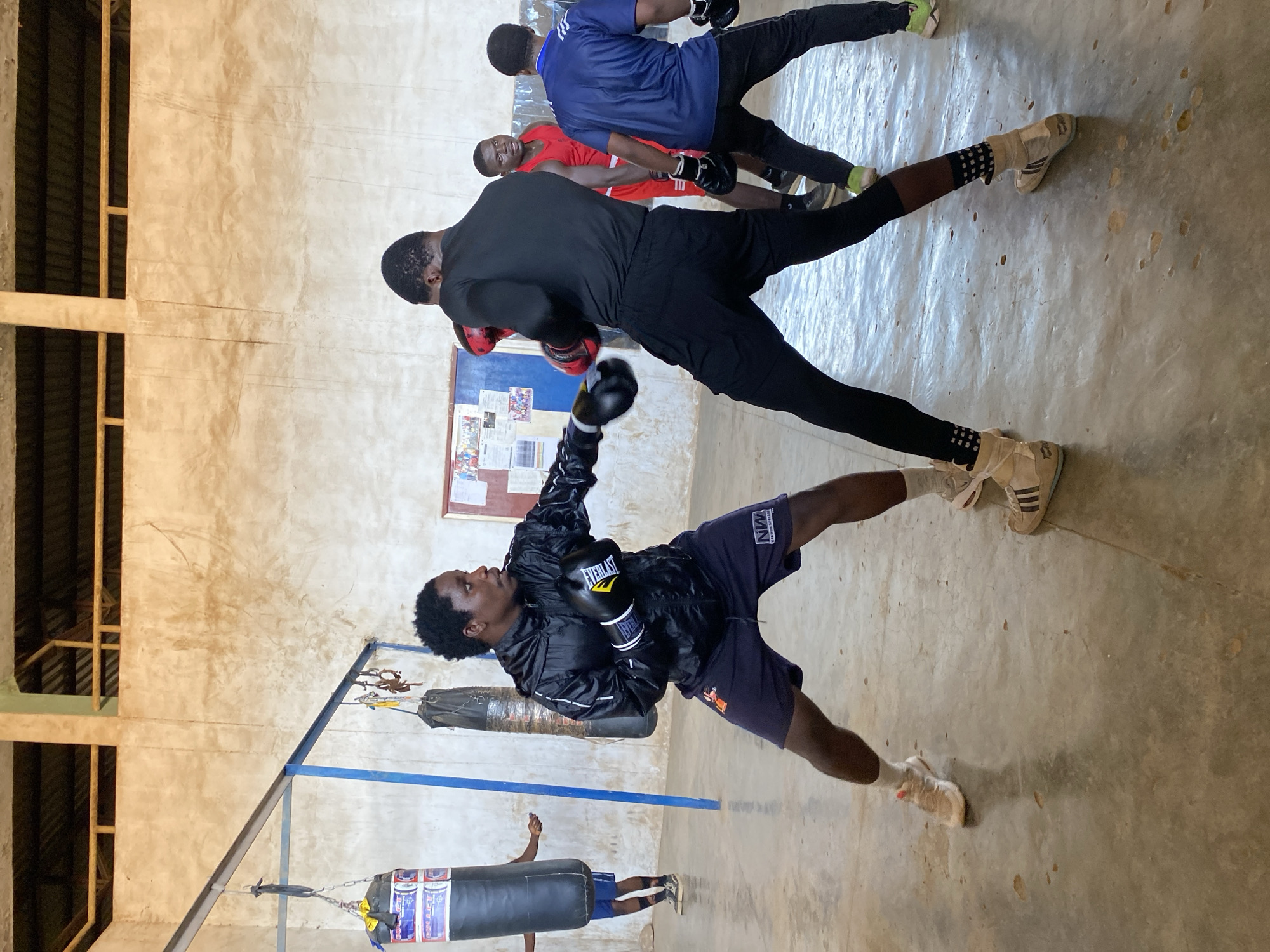 In an indoor boxing gym in Jos, Nigeria, a young man wearing a black jacket and dark blue shorts is practicing boxing. He wears Everlast boxing gloves and white sneakers with socks. He is positioned in front of a punching bag hanging from a blue metal frame. Behind him, another group of boxers are sparring: one in a black shirt and pants, one in a blue shirt, and one in a red shirt. The gym has a rustic appearance with bare walls and a simple concrete floor. A bulletin board is mounted on the wall behind the sparring boxers.