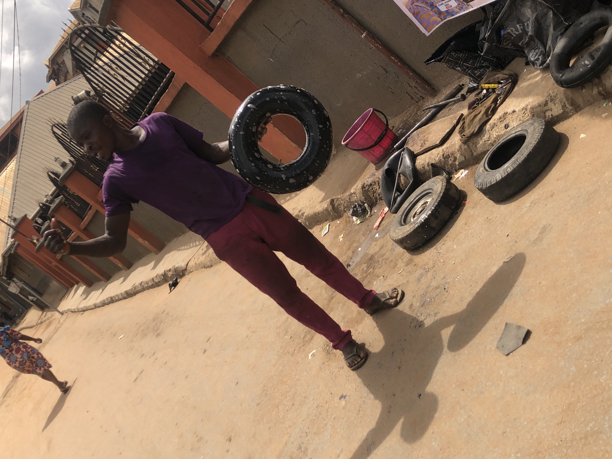 On a sunlit street in Onitsha, Nigeria, a young man stands holding a tire. He is wearing a purple shirt and maroon pants, with sandals on his feet. The ground around him is bare earth, with several tires, a red bucket, and automotive tools scattered about. A woman in a floral dress walks in the background. Buildings are visible in the background, with a metallic rooftop and an orange wall with wrought iron.