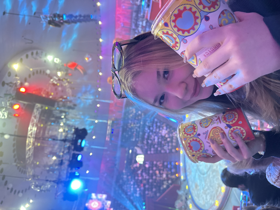 The image captures a young blonde female, approximately 18-25 years old, smiling directly at the camera while seated in the audience of a large indoor entertainment venue in Kazan, Republic of Tatarstan, Russia. She has long hair and wears black-framed eyeglasses on top of her head. Her nails feature a light-colored manicure. She holds two cylindrical, brightly patterned containers, possibly cups or popcorn tubs, featuring a folk-art design with hearts, gears, and letters; the word "КАЗАНЬ" is visibly printed vertically on the container in her right hand. The background reveals a wide, circular performance area below, illuminated with blue light patterns, surrounded by tiered seating populated by numerous indistinct spectators. The venue's high ceiling displays extensive stage lighting rigs emitting prominent red, blue, and white light, indicative of a live performance setting such as a circus or arena show. The back of another spectator's head is visible in the lower-left foreground.